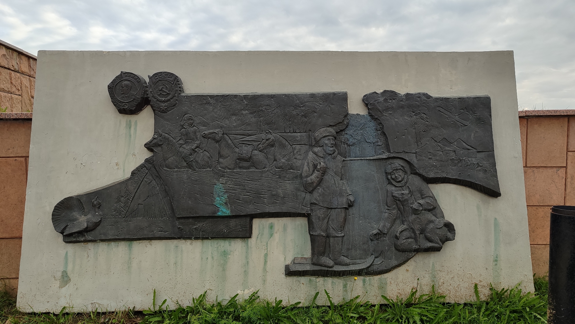 The image shows a large, dark bas-relief sculpture mounted on a light-colored, weathered wall. The sculpture depicts several scenes and figures. On the far left, a bird, possibly a turkey, is shown. To its right, there are images of trees and what appears to be a small building or structure, with an airplane in the sky above it. In the center and right sections, human figures are depicted in a historical or narrative context. Two figures are on horseback, with one leading the other, and a third figure, a man in traditional winter clothing, stands in the foreground. To the right of him, another figure in similar attire is seated. Two circular medallions, possibly emblems or portraits, are affixed to the upper left portion of the sculpture, one featuring a profile of a man and the other a coat of arms with the letters "СССР" (USSR). The sculpture is located outdoors, with greenery and grass visible at its base. A tiled wall and a black cylindrical object are present in the background to the right, suggesting an urban or park setting. The sky above is overcast with clouds. This bas-relief is likely a monument or public artwork.