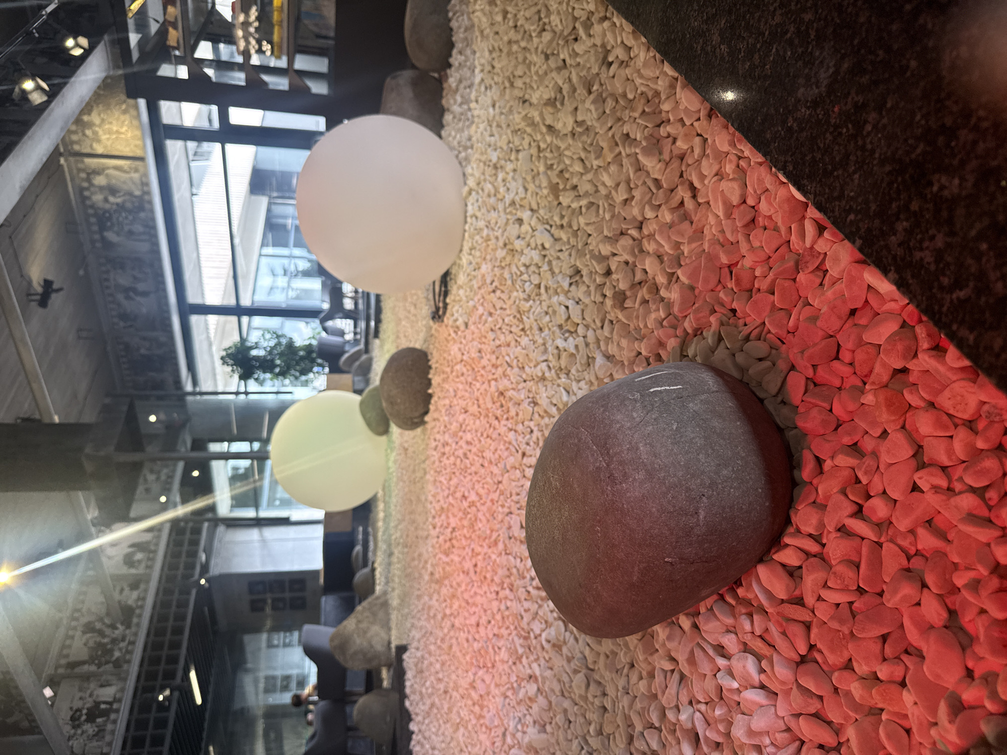 The image depicts an indoor Zen garden-like setting with a large, dark gray, smooth, oval-shaped stone prominently featured in the foreground. This stone rests on a bed of small, light-colored pebbles that are illuminated with red lighting. In the background, there are two large, spherical, glowing light fixtures, one appearing pale green and the other white. Several other stones of varying sizes are scattered throughout the pebble-covered area. The environment suggests a modern interior space, possibly a restaurant or a lounge, with elements of natural design. A beam of sunlight or artificial light streaks down from the upper left, creating a lens flare. In the far background, through large windows, is a glimpse of an urban landscape with buildings and sky, consistent with an outdoor city environment. The overall impression is one of a tranquil, artistic, and contemporary space. The location is stated as Yalta, Ukraine.