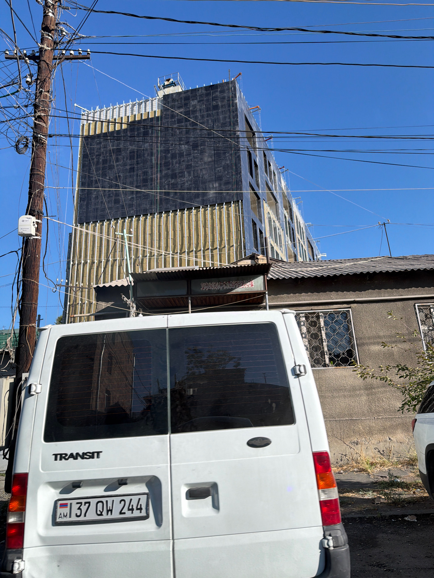 The image captures a street scene in Yerevan, Armenia, with a white Ford Transit van parked in the foreground. The van's rear is visible, displaying an Armenian license plate with the number "37 QW 244". Behind the van, a multi-story building under construction dominates the background. This building features a dark facade with vertical yellow insulation panels. Adjacent to the construction site is a smaller, older building with a corrugated metal roof and barred windows. A utility pole with numerous wires is situated to the left of the frame, further emphasizing the urban setting. The sky is clear blue, suggesting fair weather. The overall impression is one of ongoing development and a mix of old and new architecture.