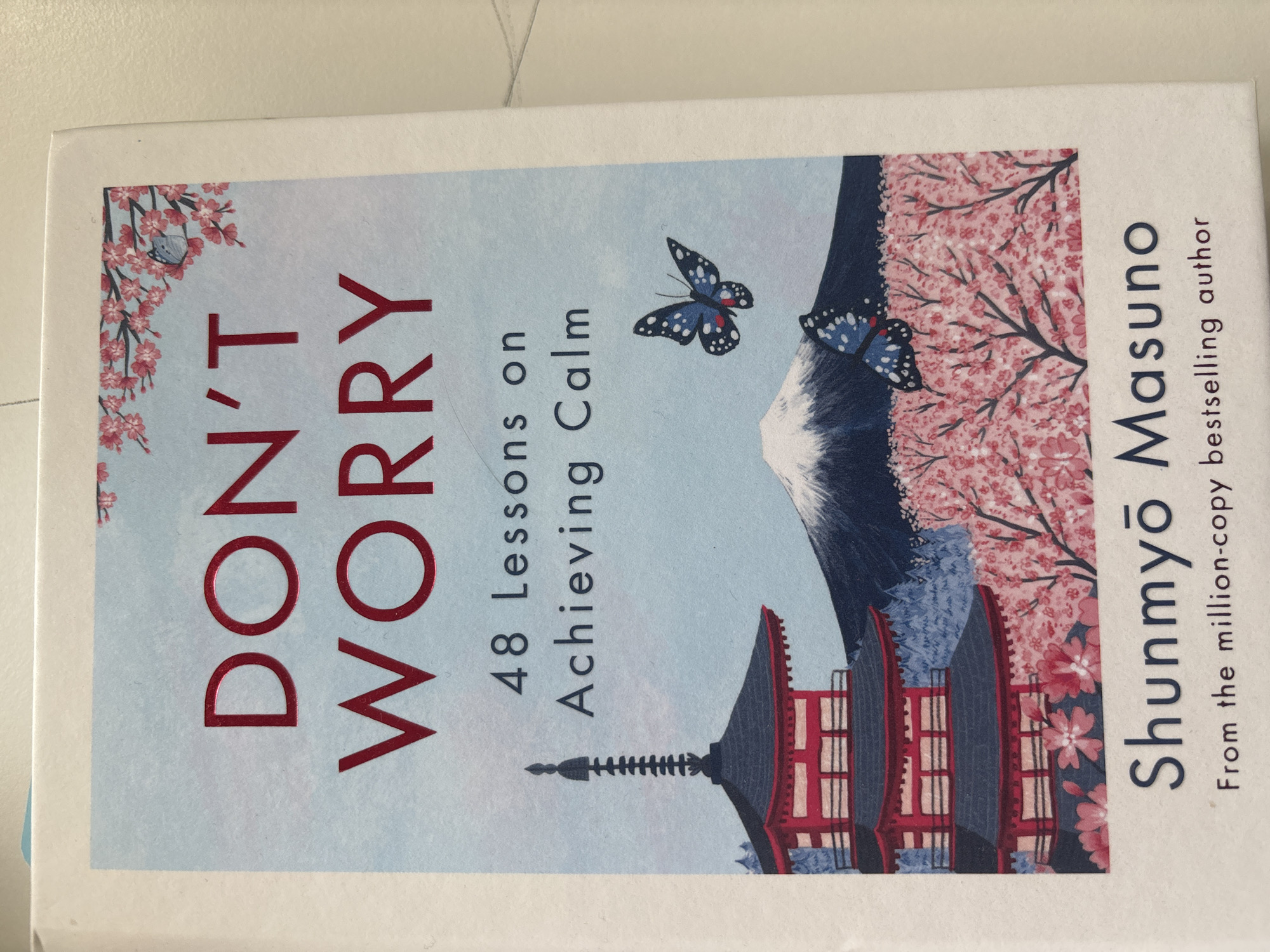 The image shows the cover of the book "DON'T WORRY 48 Lessons on Achieving Calm" by Shunmyō Masuno. The cover features an illustration with a Japanese pagoda on the left, Mount Fuji in the background, and cherry blossoms and butterflies in the foreground. The book is placed on a light-colored surface, possibly a table or desk. There are no indications of Dubai, United Arab Emirates, or any specific city in the image.