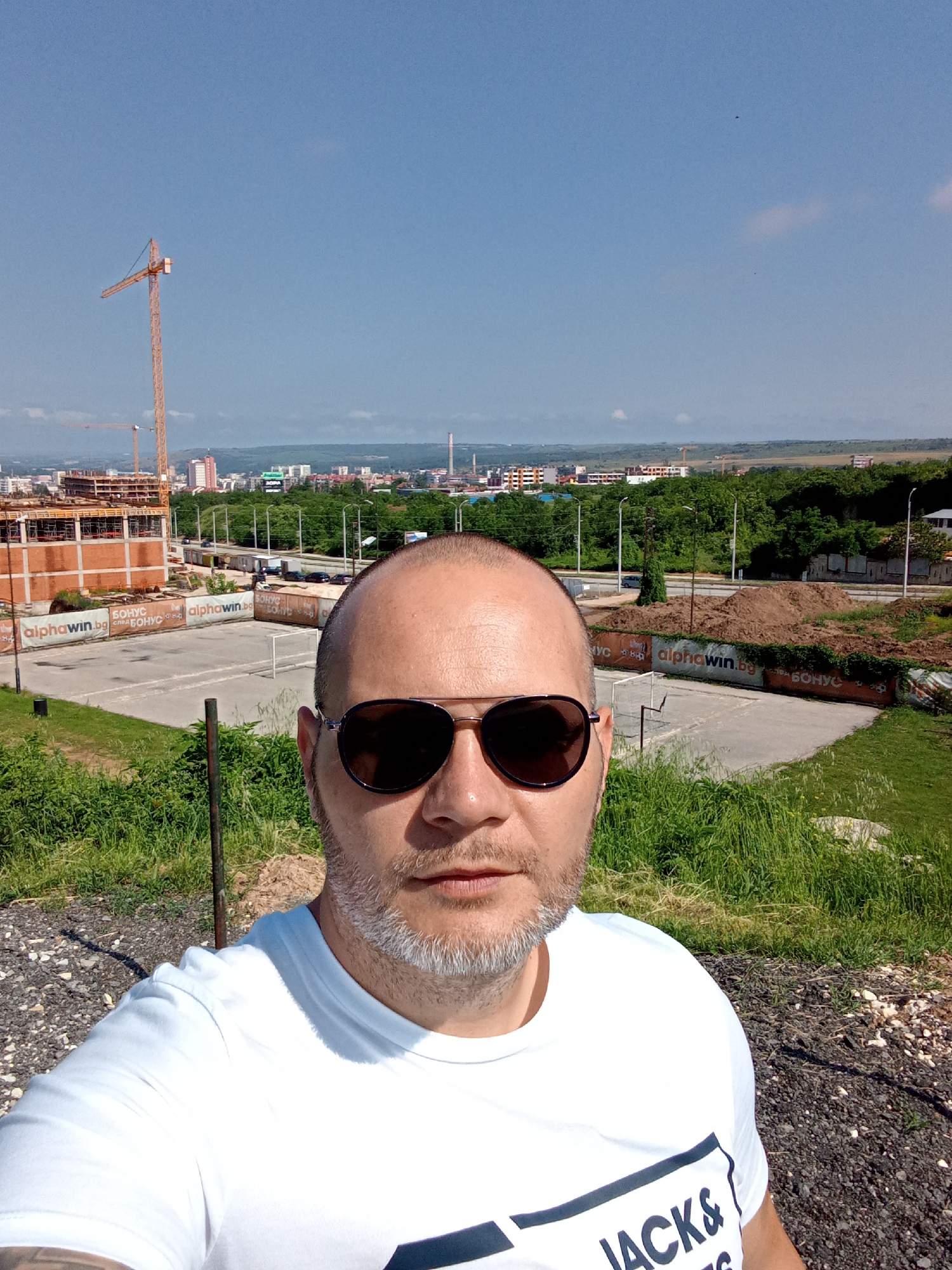 A man with a bald head, dark sunglasses, and a short grey beard is centered in the foreground, taking a selfie. He wears a white t-shirt with part of a black logo reading "JACKS &" visible on the chest.
Behind him, the scene unfolds from a high vantage point overlooking a city. To the left, there's a construction site featuring a prominent orange tower crane and a multi-story brick building under construction. Below the construction site and stretching to the right is a rough, open area with gravel, patches of green grass, and large piles of brown dirt. A small, white soccer goal sits on a paved section of this area.
A road with some vehicles runs through the mid-ground, flanked by green trees and foliage. Beyond this, a cityscape with numerous buildings of varying heights extends towards the horizon. Another smaller crane and an industrial chimney are visible in the distance. The sky above is clear and bright blue, with only a few faint clouds. Strong sunlight indicates a clear, sunny day, likely midday or early afternoon.
Several advertising banners are visible around the construction area and on fences, displaying the text "alphawin.bg" and "БОНУС" (BONUS in Bulgarian). The overall setting combines urban development with natural elements, reflecting a typical view of Pleven, Bulgaria.