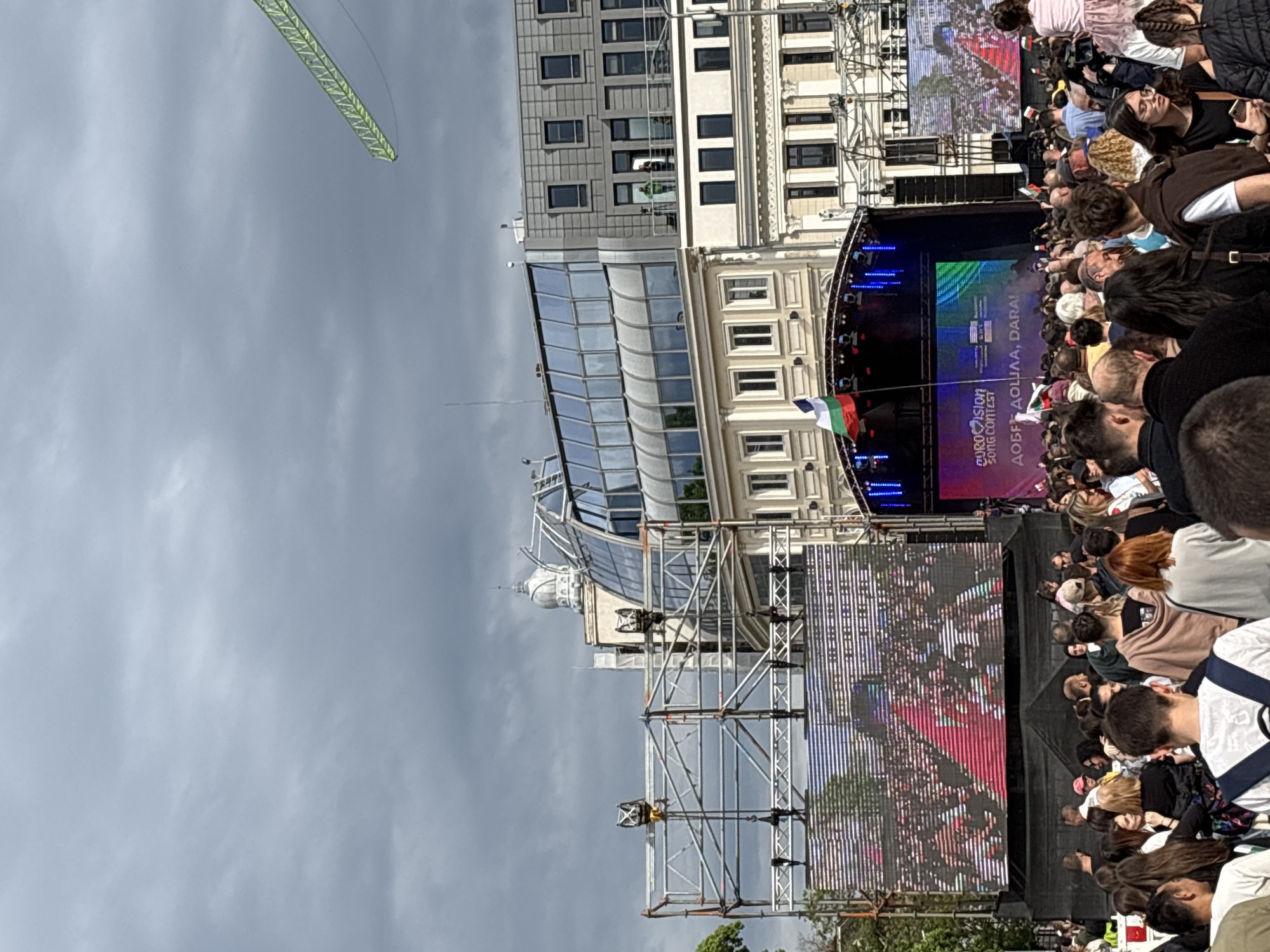 The image shows a large crowd gathered in a public square, likely in Sofia, Bulgaria, indicated by the Bulgarian flag on a stage. The event is probably related to the Eurovision Song Contest, as suggested by the event banner. There is a stage set up with large screens displaying images and text, surrounded by scaffolding. Buildings with distinct architectural styles are visible behind the stage. A crane-like structure is visible in the sky. The sky is overcast, suggesting a cloudy day. The crowd consists of many people of various ages and appearances.