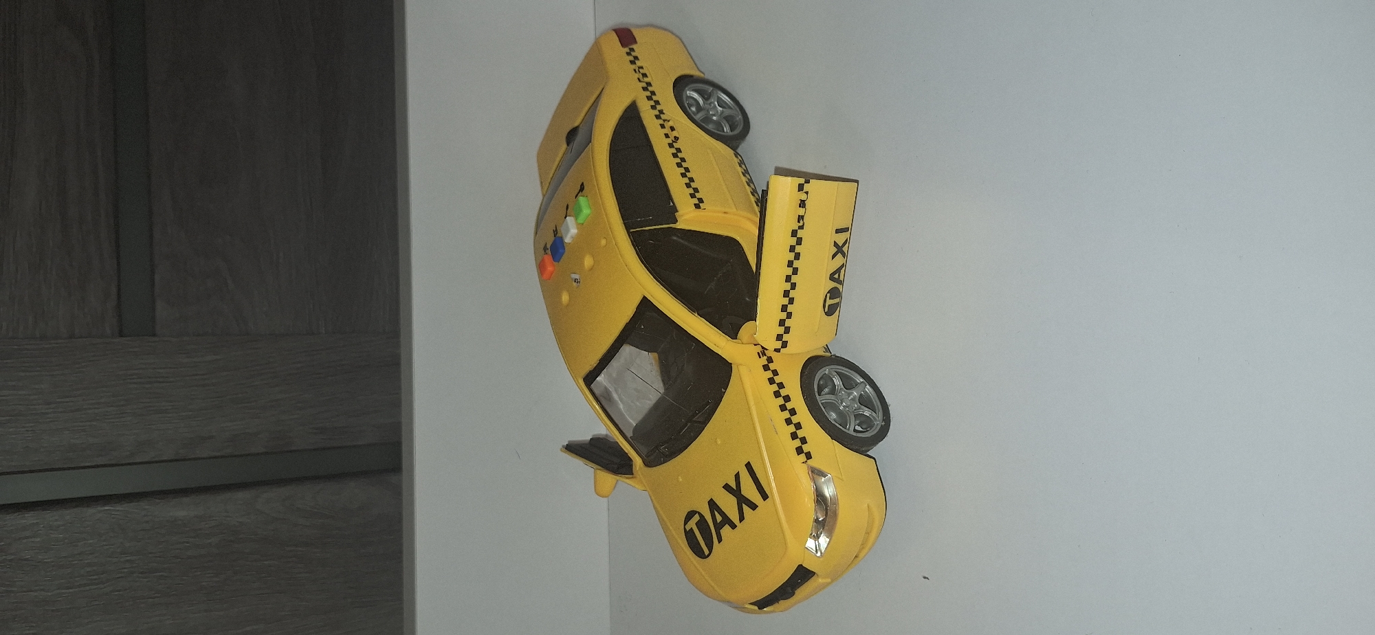 A yellow toy taxi is positioned on a white surface, with a blurred dark background of a wooden cabinet. The taxi has its driver-side door open and the passenger-side door also ajar. The word "TAXI" is visible on both the hood and the front passenger door. There are checkered black and white stripes above the front wheel wells and on the sides of the car. On the roof of the taxi, there are four colored buttons: orange, red, blue, and white. The car's wheels are silver and have five spokes. The image is taken from a slightly elevated perspective, looking down at the toy. There are no people or animals present. The scene does not provide any information to determine its location in Bila Tserkva, Ukraine.
