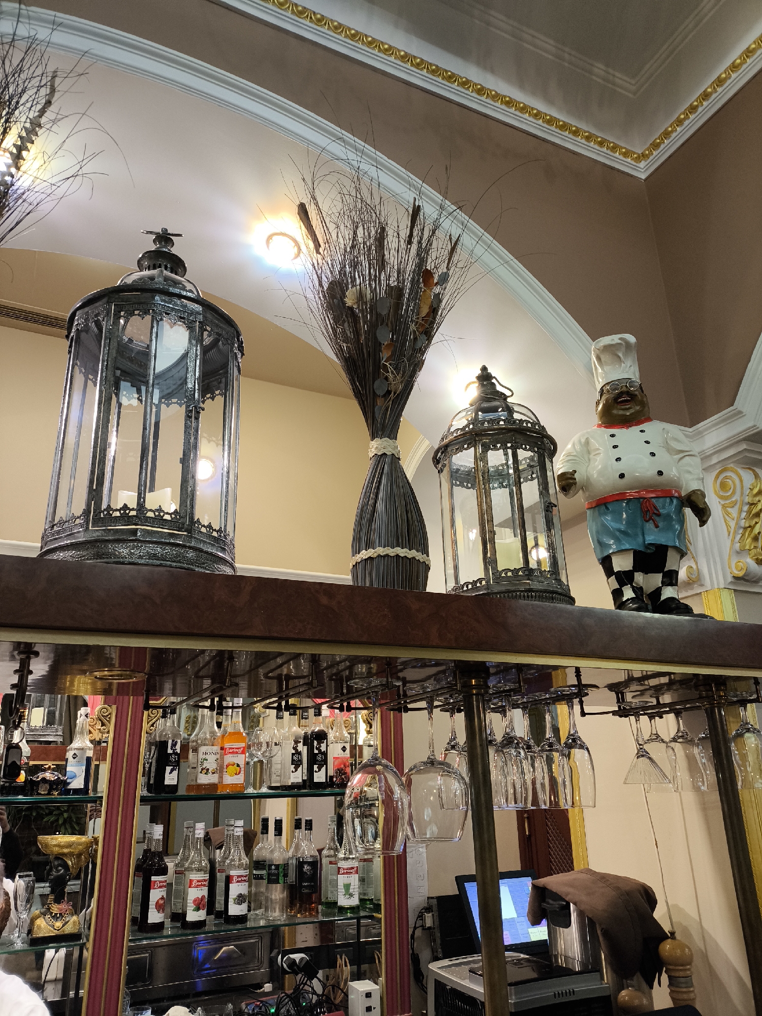 The image shows a bar area, possibly in a restaurant or hotel, featuring a shelf above the bar displaying decorative items. On the shelf, there are two ornate metal lanterns with glass panels, flanking a tall decorative vase filled with dried grasses and leaves. To the right of the rightmost lantern is a figurine of a chef with a white chef's hat and outfit, blue shorts, and checkered socks.
Below the shelf is the bar itself, stocked with numerous bottles of liquor and syrup. Wine glasses are hanging upside down from a rack above the bar. Behind the bar, there appears to be a point-of-sale (POS) system with a computer monitor and various equipment.
The walls are decorated with ornate molding and trim.
While the location is suggested to be Moscow, Russia, the image does not provide definitive visual confirmation.