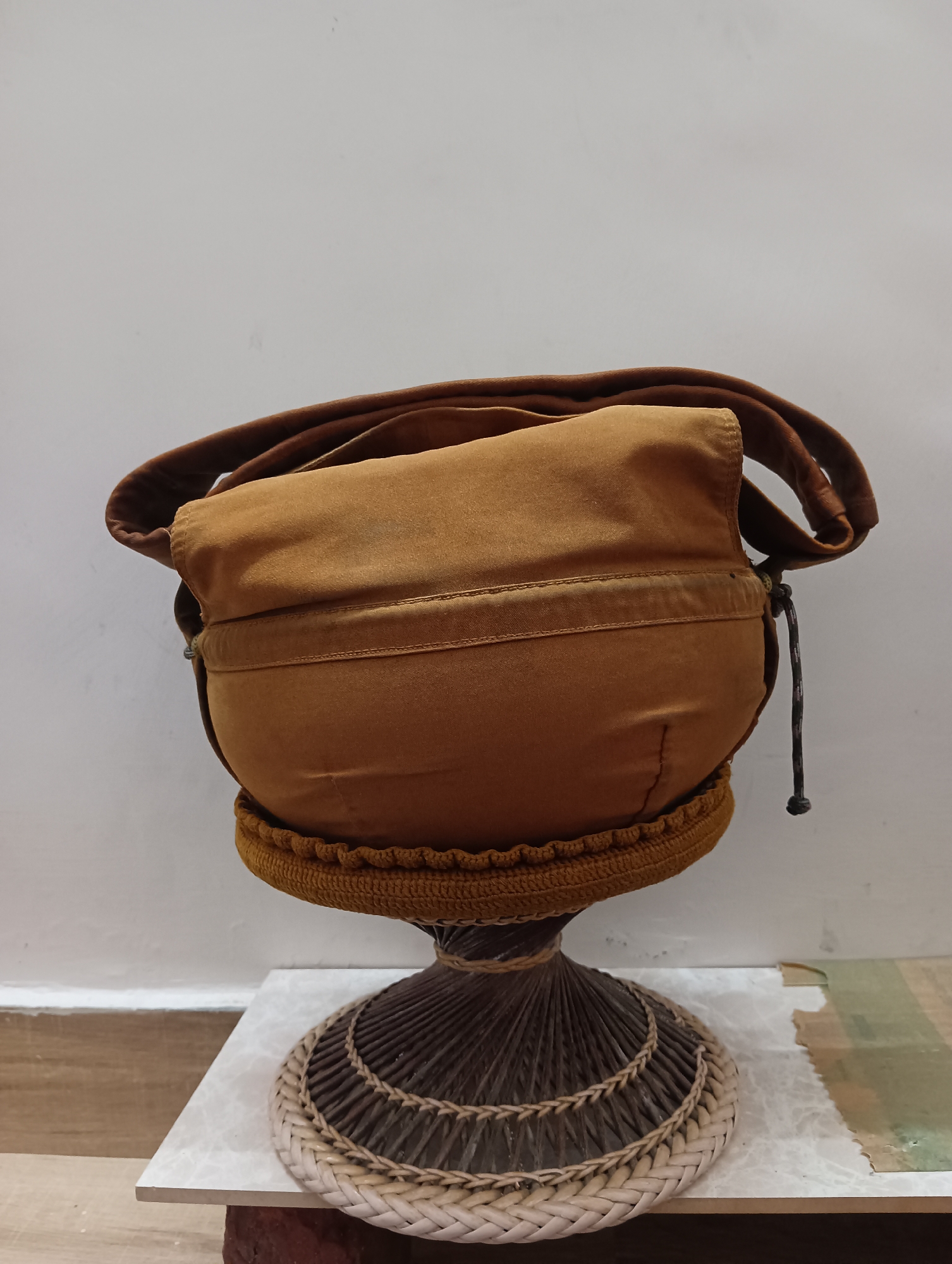 A Buddhist alms bowl, typically used by monks, is displayed on a woven stand. The bowl is made of a tan fabric and has a strap attached to its side, suggesting it can be carried. The stand is intricately woven from dark and light-colored materials, creating a decorative base. The bowl and stand are placed on a white surface, possibly a tabletop or shelf, with a plain white wall in the background. No people are present in the image, and there are no discernible elements that would suggest a specific location in Manali, India. The scene is static and appears to be a studio or home setting for display.
