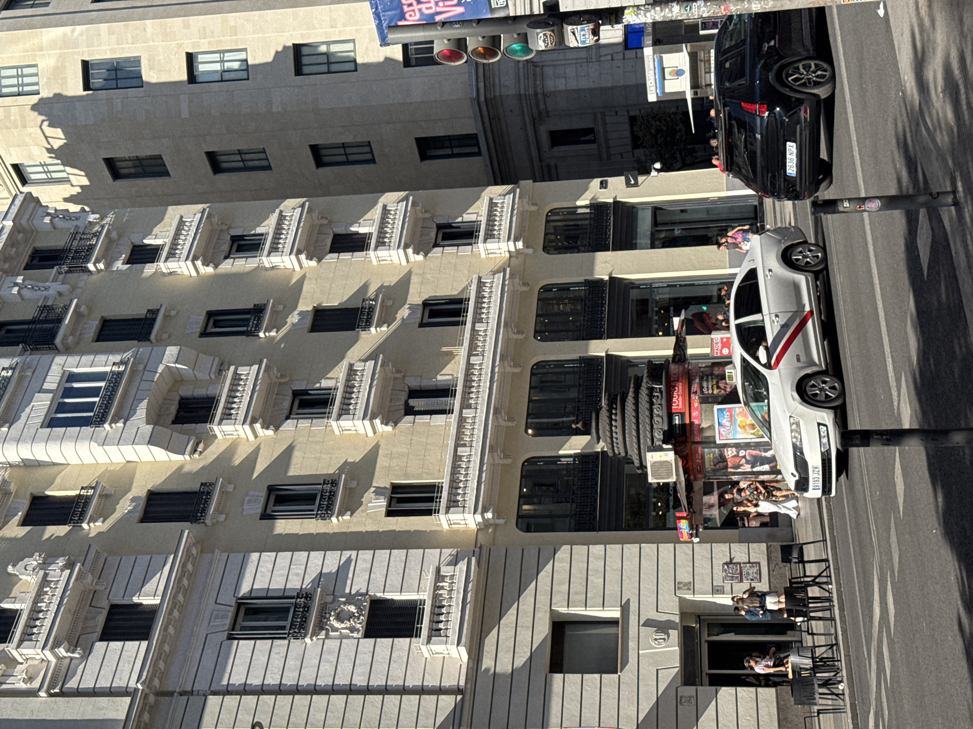 In Madrid, Spain, on a sunny day, a traffic light signals red for a black SUV and a white taxi. The taxi, with license plate 9193 JZV, is in the foreground on the right, facing left. The black SUV, with license plate 2636 NPX, is behind it, also facing left. Both vehicles are stopped at a crosswalk in front of buildings with traditional Spanish architecture. The buildings are tall, with multiple stories featuring balconies with ornate railings and large windows. Some windows are open, and some have shutters. The facade of the buildings vary in color from pale yellow to a lighter stone color. On the left side of the image, people are standing outside a cafe with tables and chairs. Further down the street, other pedestrians are walking and interacting. A newsstand or kiosk is visible, with posters advertising various events. The sky is clear and blue.