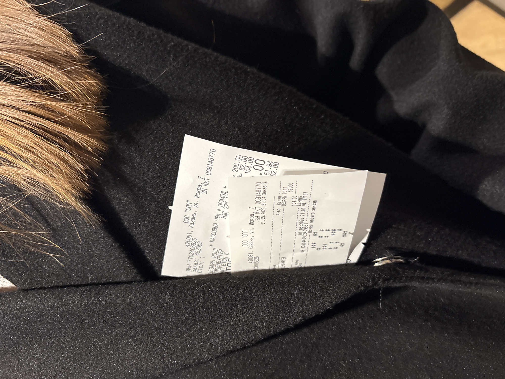 An angled overhead shot captures a white paper receipt tucked into or resting on a dark, possibly black, piece of clothing, which appears to be a coat or jacket. The clothing has a brown, shaggy fur trim visible on the left side of the frame. The fabric of the garment shows a textured, slightly uneven surface.
The crumpled receipt is a cash register check from "ООО 'СПП'" (OOO "SPP"), located at "420081, Казань, ул. Искра, 7" (420081, Kazan, Iskra Street, 7), placing the transaction in Kazan, Russia. The INN (taxpayer ID) 7703406825 is visible. The check indicates "ПРИХОД" (ARRIVAL) and "НДС 22%" (VAT 22%). The transaction took place on "01.05.2026" (May 1, 2026) at "21:04" and "21:08," indicating late evening.
Purchased items listed include "ЦЕЗАРЬ РОЛЛ" (Caesar Roll) for 82.00 and "КЕНБУРГЕР" (Kenburger) for 104.00. "МАГГЕТСЫ 6" (Nuggets 6) is also partly visible. Some prices on the receipt include 206.00, 82.00, 104.00, 51.94, and 92.00. An "Заказ: 452369" (Order: 452369) and "Стол: 1" (Table: 1) are also readable. The cash register number is "ЗН ККТ 009148770". The overall scene suggests a recent purchase from a restaurant or cafe, with the receipt left on an item of clothing.