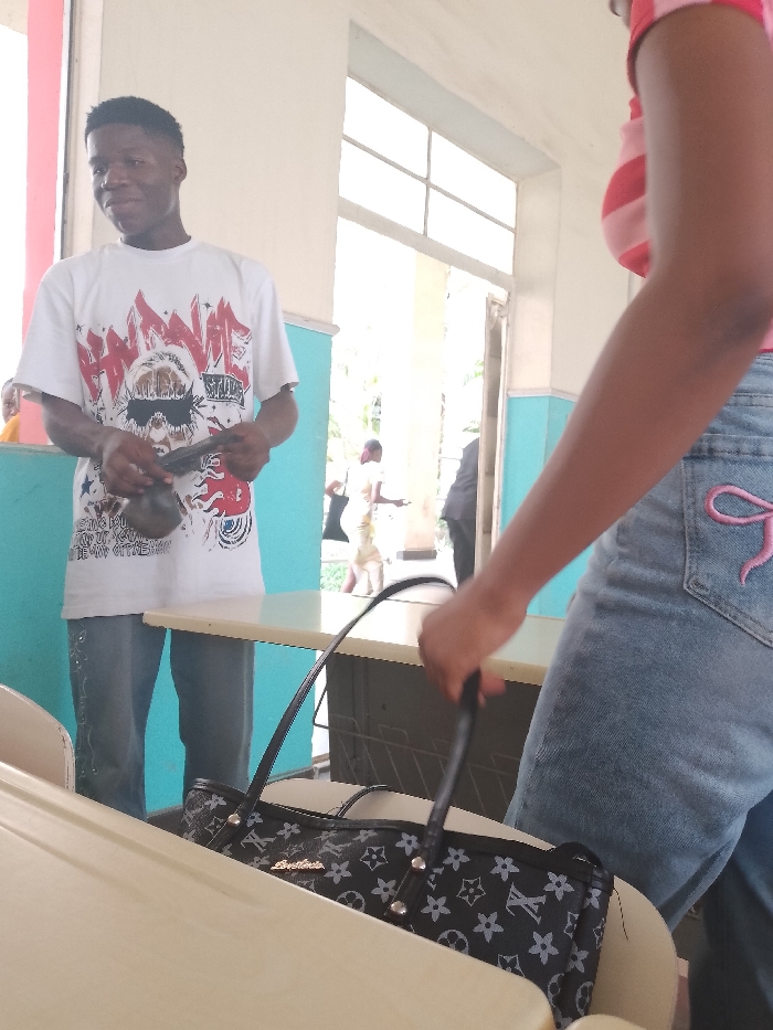 A medium shot captures a man in a white graphic tee and blue jeans, holding a dark object. He is standing to the left of a light-colored table. To the right, a woman in a striped pink shirt and blue jeans, adorned with a pink bow on the pocket, holds a black handbag on the same table. In the background, an open doorway reveals figures in motion. The location is identified as Umueshi, Nigeria.