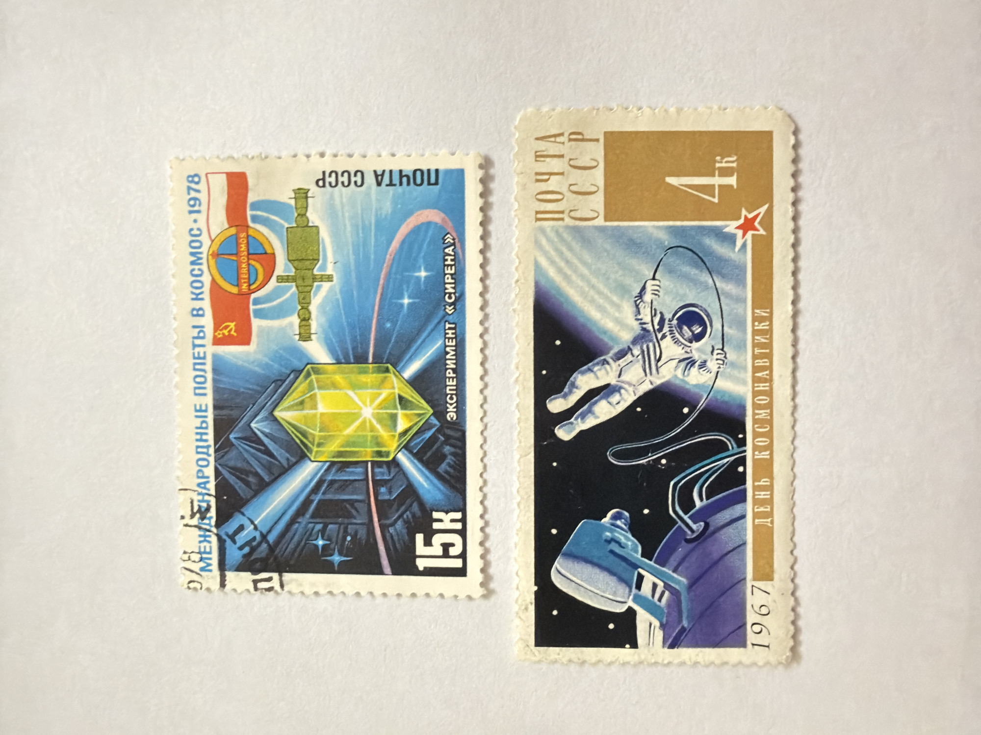 The image shows two Soviet Union postage stamps. The stamp on the left features a satellite with a yellow, dodecahedral prism at its center. The stamp is blue and black, with the words "МЕЖДУНАРОДНЫЕ ПОЛЕТЫ В КОСМОС-1978" (International flights in space - 1978) written in Russian, along with "ЭКСПЕРИМЕНТ «СИРЕНА»" (Experiment "Sirena") and "15к" (15 kopeks). The logo of INTERKOSMOS is in the upper left corner. The stamp on the right is of a cosmonaut in space, skipping rope near a space station. It is printed in blue, black, white, and gold. The stamp includes the words "ДЕНЬ КОСМОНАВТИКИ" (Cosmonautics Day) and "1967", along with "4K" (4 kopeks) and "ПОЧТА CCCP" (Post of the USSR). The location of Izhevsk, Russia is not relevant to the image content.