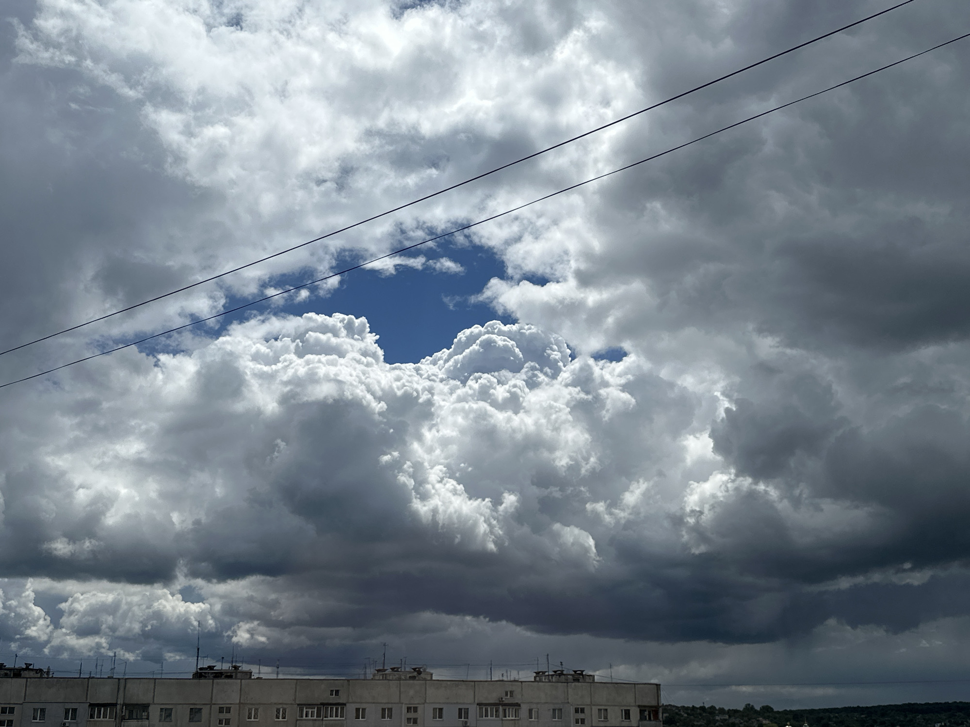 The image shows a building in the foreground, with a cloudy sky overhead. The building is a light grey color and has several windows. There are some objects on top of the building, possibly antennas or ventilation units. The sky is filled with a mix of dark grey and white clouds, with a patch of blue sky visible in the center. There are two horizontal power lines visible across the sky. The location is a city of Unknown, Unknown.