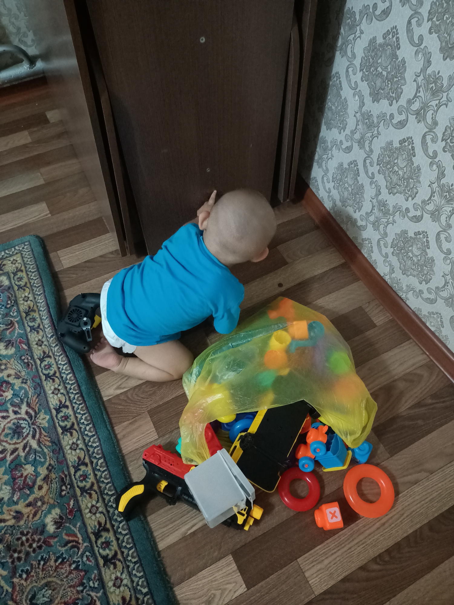 In a room with wood-paneled walls and patterned wallpaper, a toddler wearing a blue t-shirt and a diaper kneels on a wooden floor. A colorful, patterned rug is visible to the left of the toddler. The child is partially obscured by a large, bright yellow plastic bag filled with various toys, including colorful plastic blocks and a toy gun. Among the scattered toys are red and orange rings, a small orange block with an "X" on it, and parts of what appear to be construction toys. To the left of the toddler, near the rug, is a black toy with yellow accents, possibly a vacuum cleaner. The scene suggests a child playing indoors. The lighting appears to be artificial, indicating it is likely daytime or evening. No text is visible in the image.