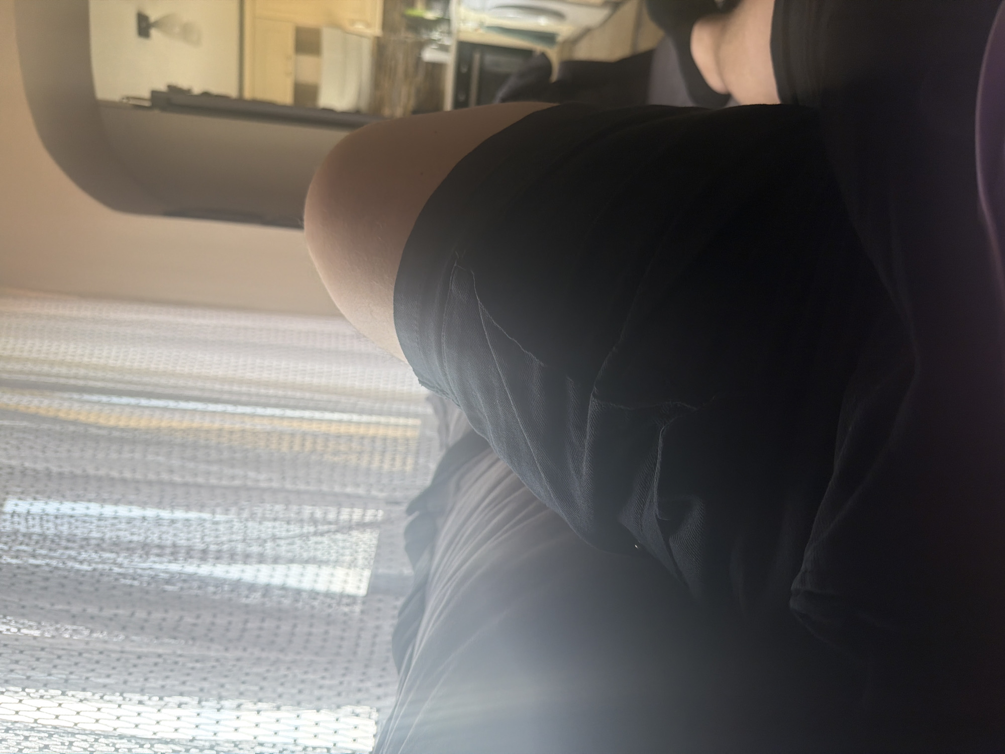 This image, oriented vertically as if rotated 90 degrees clockwise, depicts an indoor scene with a close-up on a person's lower body. The main subject is a person, likely lying down or sitting in a relaxed position, whose upper thighs and hip area are visible in the foreground and mid-ground. They are wearing black shorts or leggings, and their skin is light-toned. To the right, additional dark fabric, possibly purple, is present, which could be clothing or bedding.
In the left portion of the image, a prominent light-colored, textured fabric, resembling a curtain or window blind, suggests the presence of a window from which bright daylight is entering. This indicates the scene takes place during the daytime.
The background, which appears inverted due to the image's orientation, shows an indoor environment. Faintly visible elements suggest a room, possibly a kitchen or another living space, with light-colored furniture, shelves, or architectural features. There are no specific activities taking place other than the person resting. No text is visible in the image. The setting is an indoor space, possibly in Shumen, Bulgaria, as per the provided context.