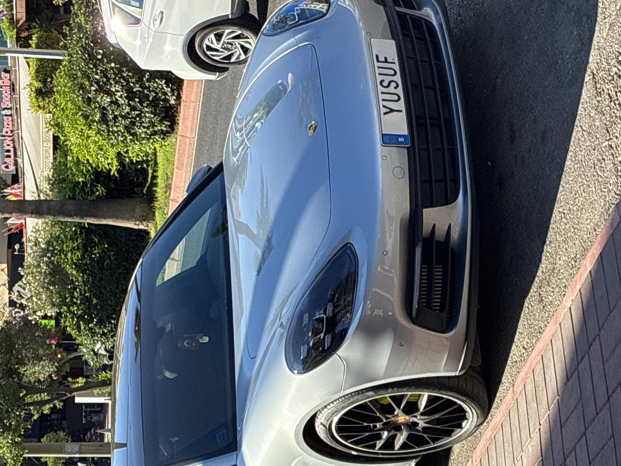 A silver Porsche Panamera is parked on a city street in Alanya, Turkey, bathed in bright sunlight. The car's license plate reads "YUSUF" and features the Swedish flag emblem. In the background, a white SUV is also visible, along with a bus stop sign and lush green bushes. Further back, a restaurant called "GALLION Pizza & Snack Bar" is identifiable by its signage. The car is parked on the right side of the road, with a sidewalk made of patterned bricks in the foreground. The overall scene depicts a typical urban environment on a sunny day in Alanya.