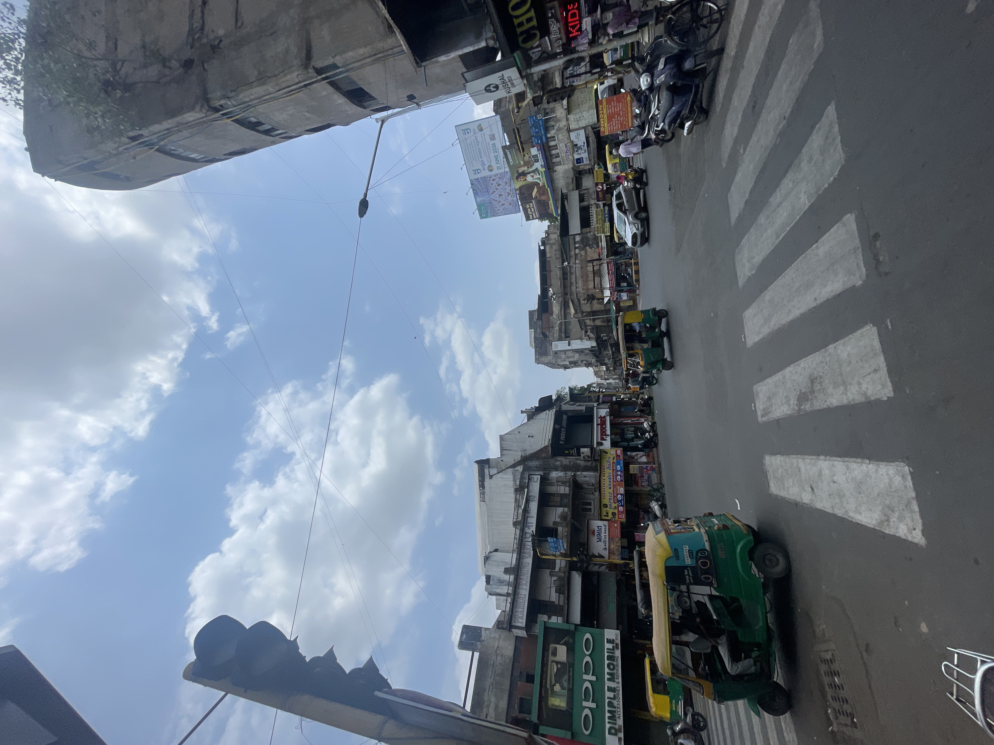 The image captures a street intersection in Ahmedabad, India, under a partly cloudy sky. A traffic light is visible on the left, casting a shadow. In the foreground, a green and yellow auto-rickshaw is stopped at a pedestrian crossing. The street is paved with asphalt, and white stripes demarcate the pedestrian crossing. Various shops and buildings line the sides of the street, displaying signage for businesses like "OPPO" and "DIMPLE MOBILE." Several other vehicles, including cars and motorcycles, are present on the road, with some people walking on the sidewalks. A tall, somewhat dilapidated concrete building with visible windows stands on the right side of the intersection, and a street lamp is mounted on a pole nearby. Billboards with advertisements are also visible on some of the buildings. The scene depicts a typical urban street scene with moderate traffic and commercial activity.