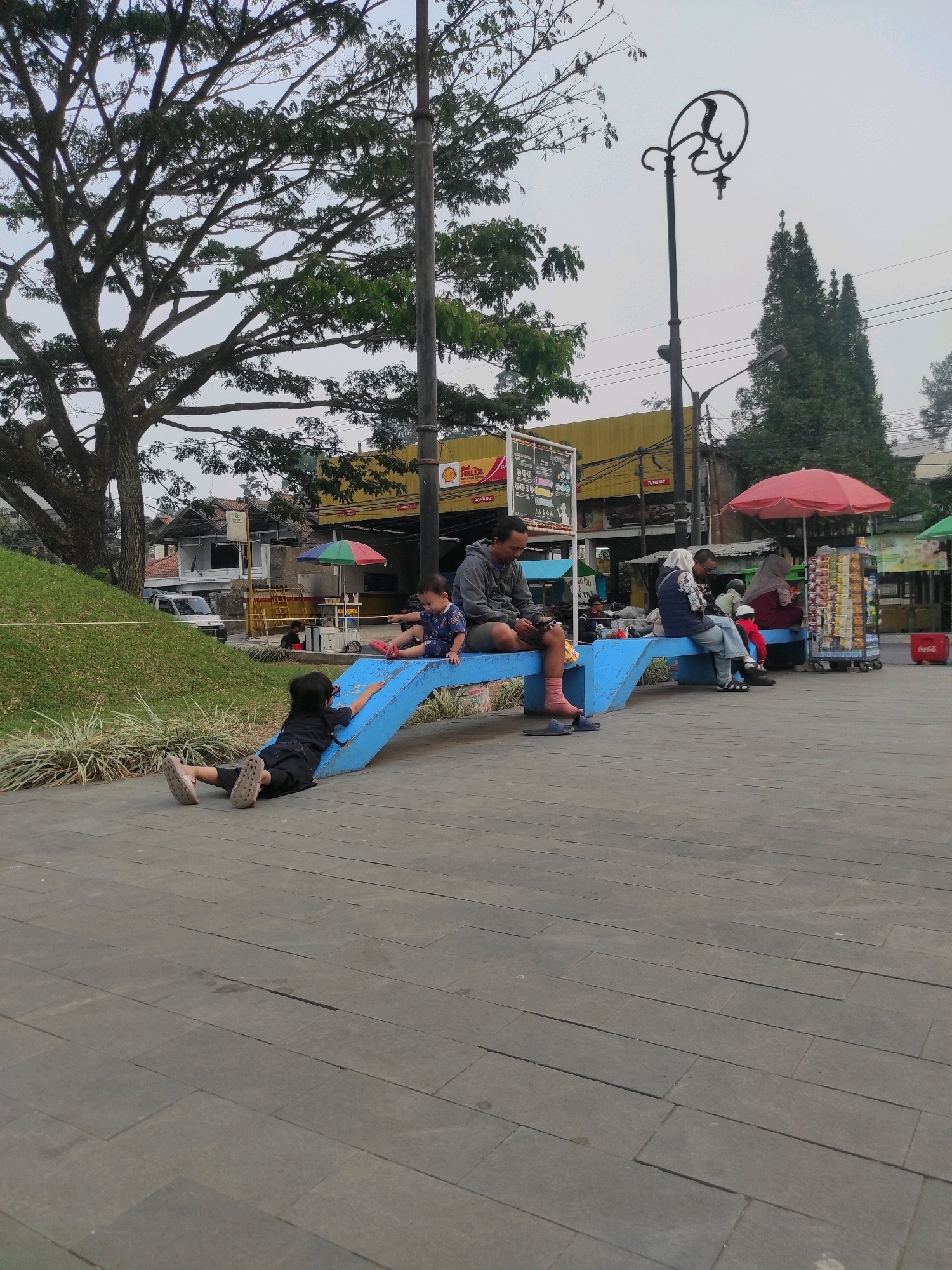 In Ciburuy, Indonesia, a paved area features a blue, wave-like bench. A man sits on the bench, looking at his phone, with a child beside him. Another child is lying on the lower section of the bench. Several other people are seated on the bench. A street vendor's cart with colorful snacks is visible, along with red and green umbrellas. In the background, there are buildings, trees, and a lamppost. The ground is paved with large, gray rectangular tiles.