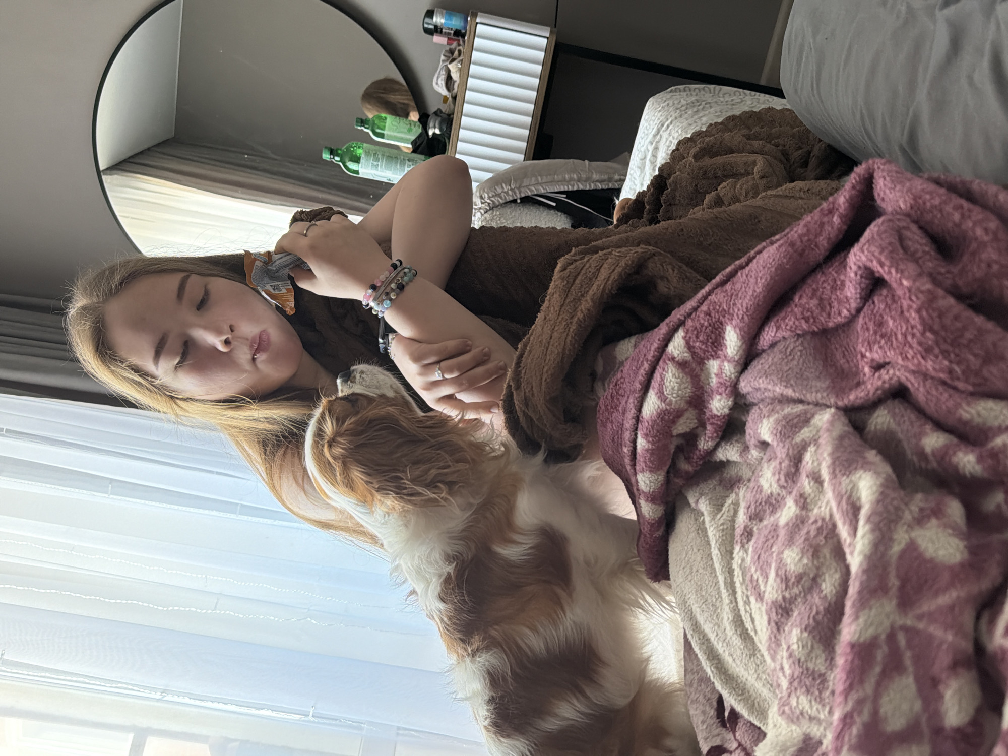 The scene depicts an indoor setting, likely a bedroom, in the city of Chaikovsky, Russia. A young woman with light brown hair is seated on a bed, covered by a brown blanket and a pink and white patterned fleece blanket. She is holding an open, metallic-orange snack bag to her mouth, actively eating from it. Her left wrist displays multiple beaded bracelets, and a ring is visible on her right ring finger. To her immediate left, a white and brown Cavalier King Charles Spaniel dog stands on the bed, looking upward towards the woman's face and the snack bag, with its attention directed at the food. The background features a window on the left, covered by sheer white curtains. On the right, a large round mirror with a black frame reflects a portion of the room, including a white dresser with a ribbed front, upon which two green bottles and several small cosmetic containers are arranged. The room walls are a neutral grey.