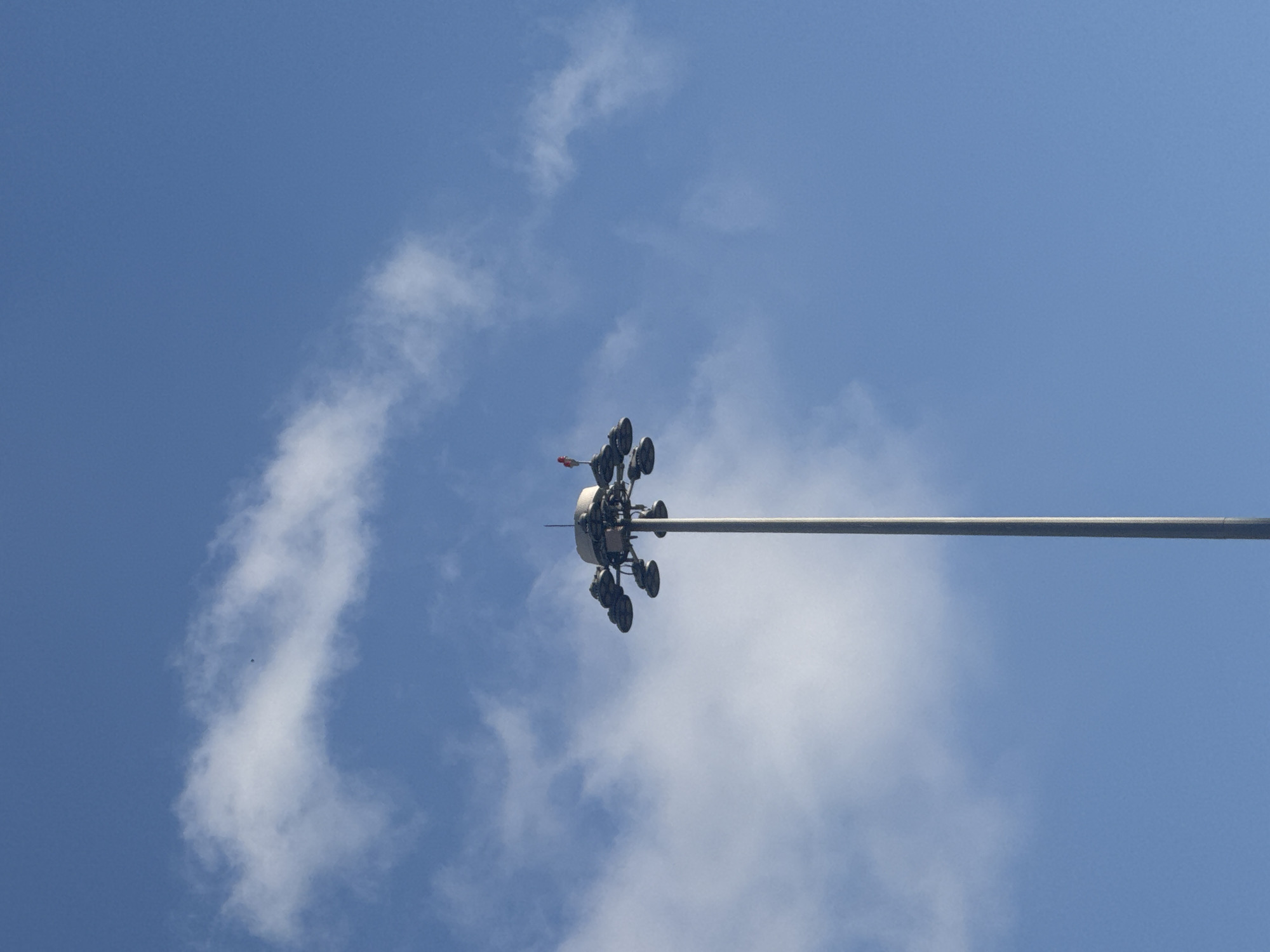 The image shows a close-up view from below of a tall pole topped with a cluster of lights, possibly a stadium or street light. The pole extends diagonally across the frame from the lower right to the upper left. The light fixture at the top is circular and complex, with multiple spotlights arranged around it. A small, reddish-orange object, possibly a flag or marker, is attached to the very top of the fixture. The sky behind the pole is a clear, bright blue with scattered white clouds. The clouds are wispy and appear to be high in the atmosphere, suggesting it is daytime, likely late morning or afternoon. The overall scene is dominated by the sky and the imposing structure of the light pole. There is no indication of people or any specific event taking place. The location context of Tel-Aviv, Israel, does not provide any visually discernible clues within the image itself. There is no visible text.