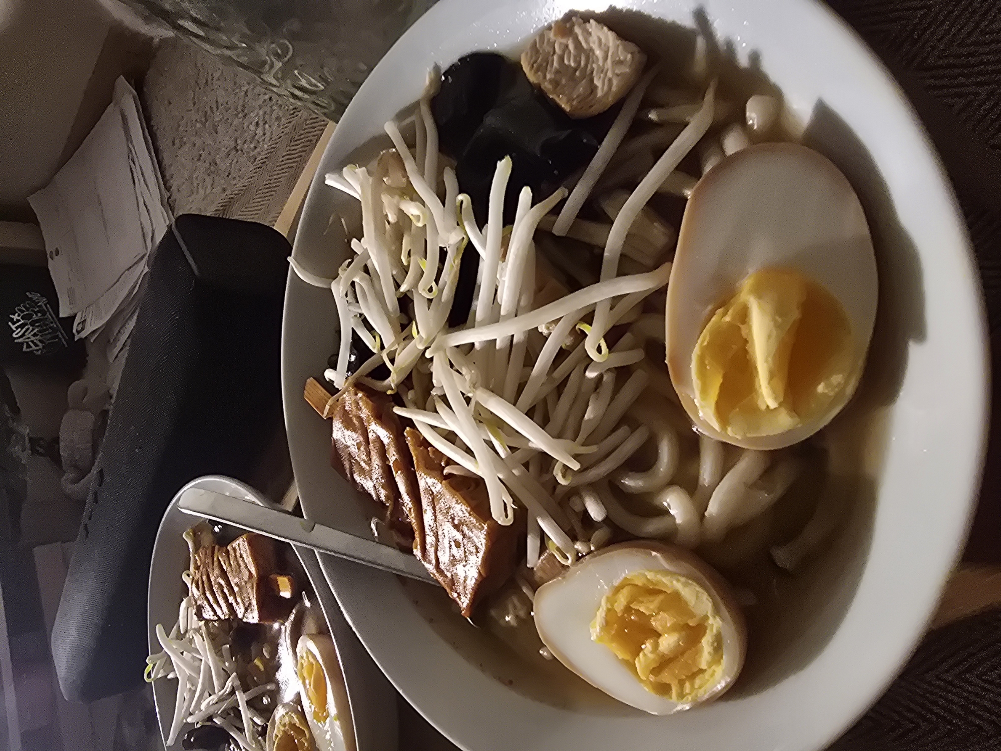 A close-up shot shows two bowls of ramen on a surface with what seems to be cushions or pillows to the side. The ramen bowls contain noodles, halved boiled eggs with cooked yolks, bean sprouts, dark-colored mushrooms, and pieces of what appears to be braised pork belly or similar meat. A pair of metal tongs rest on one of the smaller bowls. The lighting suggests an indoor setting, possibly a home environment, but there's no direct evidence of the location being in Aubervilliers, France.