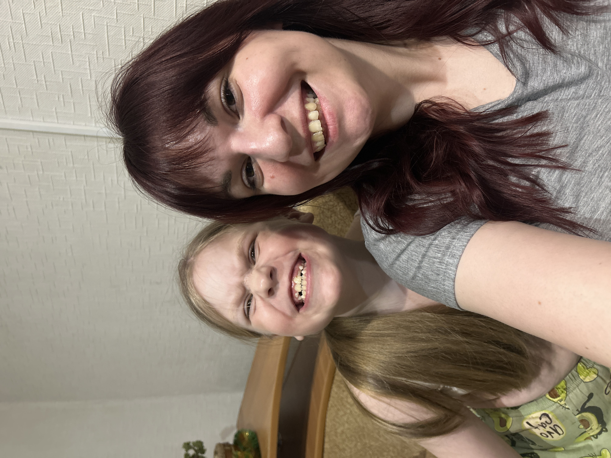Two females are posing for a selfie. One female has dark, auburn-colored hair and is wearing a gray t-shirt. The other female has blonde hair in a ponytail and is wearing green pajama pants with a pattern of avocados and black cats. The location appears to be indoors, potentially a residence. No information is available to confirm that the photo was taken in Фоки, Russia.