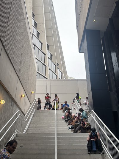 The image depicts a group of people gathered on a wide outdoor staircase situated between two tall buildings. The staircase is concrete with metal railings, leading up to a platform where more people are congregated. The architecture of the buildings suggests a modern, possibly Brutalist, style.
Many of the individuals on the stairs are seated, while a few stand. Their clothing is varied, with some wearing casual attire and others appearing to be in costumes or cosplay outfits. The presence of individuals in costumes may indicate that the location is near a convention or event space.
The photo was likely taken in downtown Atlanta, Georgia. The architectural style of the buildings in the background, and the crowd on the stairs, is consistent with locations such as the Georgia World Congress Center (GWCC) or the Hyatt Regency hotel. The people in the photo are likely gathered for a convention or event hosted in the area.