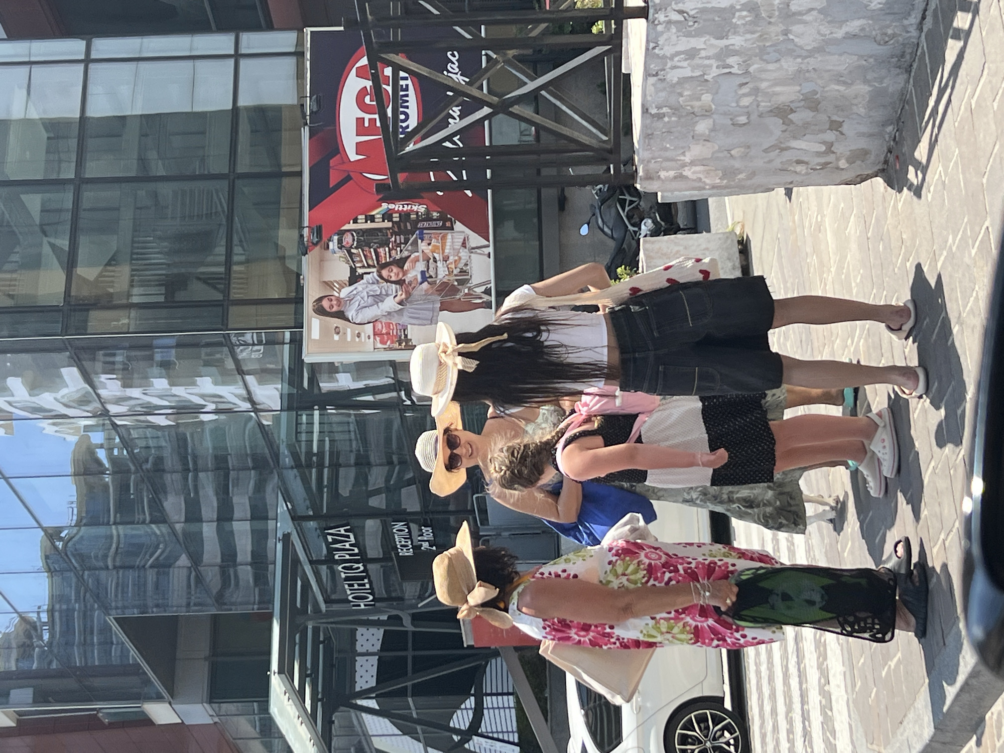 In Budva, Montenegro, a group of four females are gathered on a pedestrian walkway in front of a modern building with a glass facade, identified by signage as "HOTEL TQ PLAZA". The building's reflection shows construction or renovation work. A large advertisement featuring a woman and child in a shopping cart, alongside the text "MEGA ROMEI", is visible on the building. One woman, wearing a floral dress and a wide-brimmed hat, carries a large tote bag and a green mesh bag. Another woman, also in a wide-brimmed hat, is smiling and wearing a sleeveless top. A young girl with braided hair, wearing a black and white patterned dress, stands between them. The fourth woman, seen from behind, wears a white crop top and dark denim shorts, carrying a tote bag adorned with red hearts. A white car is parked to the left, and a motorcycle is partially visible behind a black metal railing. The ground is paved with bricks, and the lighting indicates it is daytime.