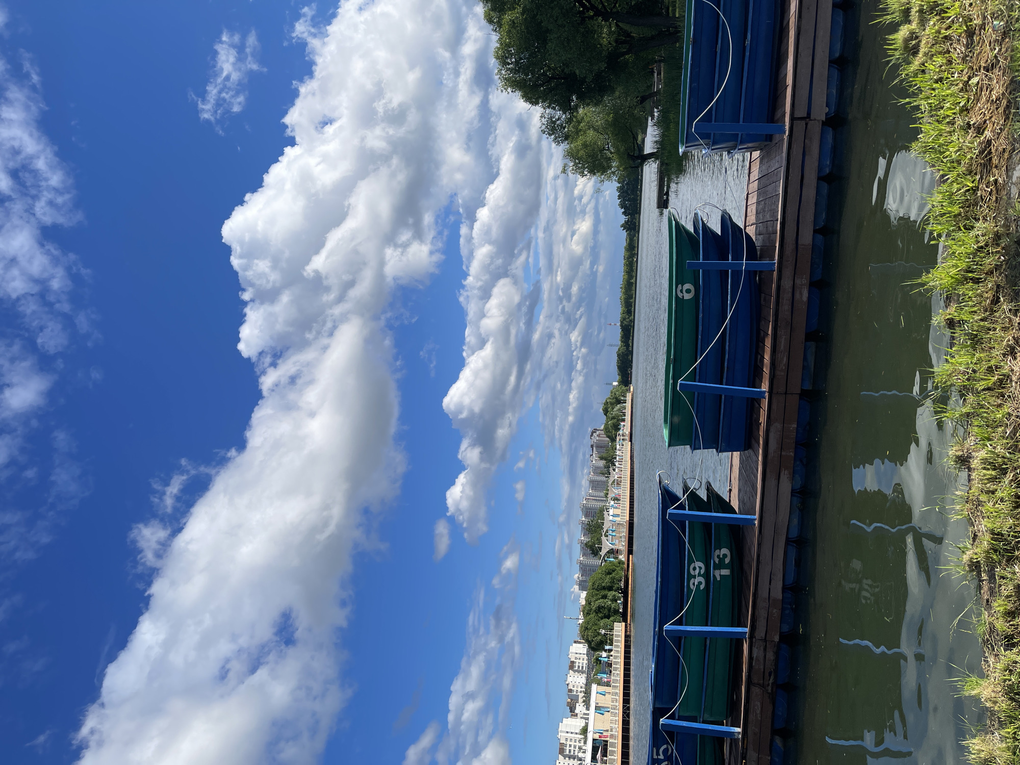 The image depicts a scenic waterfront area with several boats docked on a pier. The scene is set against a backdrop of a blue sky with fluffy white clouds. In the distance, there are buildings that suggest an urban environment. The pier itself is constructed of wood and floats on blue pontoons. Stacked on the pier are several boats, some green and some blue, each with numbers visible. One green boat has the number "6" prominently displayed. Another green boat, numbered "39", is stacked beneath a blue boat, and the number "13" is visible on a blue boat below that. The water in the foreground is calm and reflects the sky and the surrounding environment. There is also a grassy embankment in the immediate foreground.
This scene is identifiable as being located in the Zaryadye Park in Moscow, Russia. Specifically, it is the area near the Moskva River within the park, where visitors can rent boats. The buildings in the background are part of the Moscow skyline and infrastructure surrounding the park. The overall atmosphere is peaceful and recreational, typical of a public park on a clear day. There are no people visible in the image, and the boats appear to be stationary and ready for use.