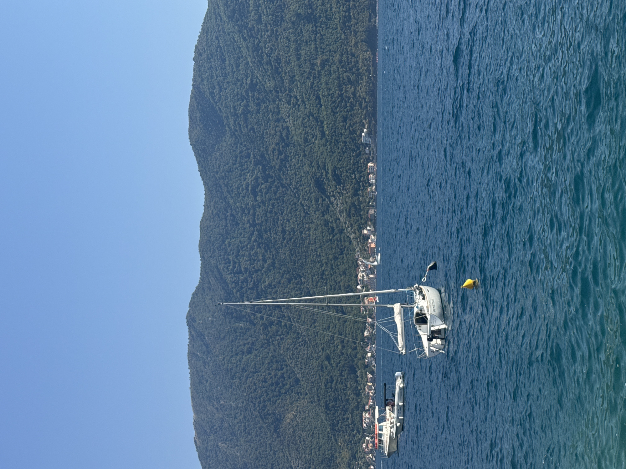 The image depicts a serene coastal scene in Perast, Montenegro, under a clear blue sky. In the foreground, a white sailboat with a mast stands anchored in the deep blue water. The boat's name, "BENETEAU," is visible on its hull. A bright yellow buoy floats nearby. Further in the distance, another white motorboat with passengers is anchored. The shoreline is lined with quaint, red-roofed buildings, characteristic of Perast's historic architecture, nestled at the foot of a large, lush green mountain. The mountain itself is densely covered with trees, indicating a natural, undeveloped landscape. The water exhibits gentle ripples, suggesting a calm day. The overall atmosphere is tranquil and picturesque, capturing the essence of a Mediterranean coastal town.