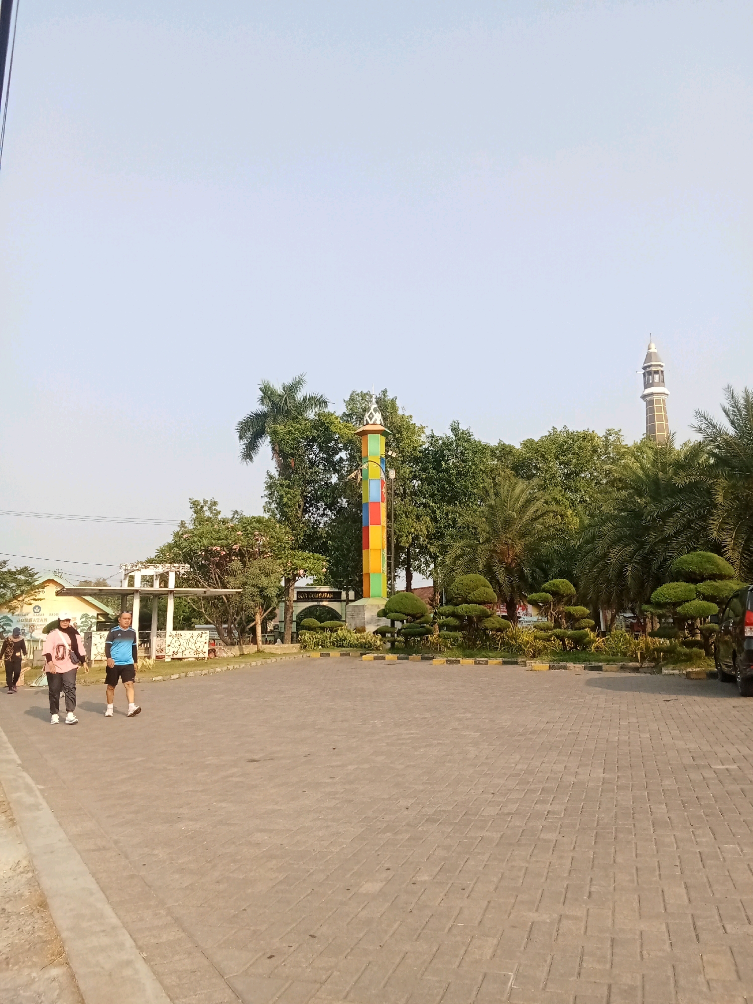 The image depicts an outdoor scene in Kaliwungu, Indonesia, likely a public park or recreational area. In the foreground, a paved walkway made of light-colored rectangular bricks is visible, leading into a larger paved area. Several people are walking on the walkway. On the left, two individuals are walking from left to right, one wearing a pink shirt with the number "0" and a white head covering, and the other wearing a blue t-shirt and dark shorts. Further back, another person in dark clothing is walking.
In the background, a tall, colorful, rectangular monument with a pattern of colored squares stands prominently. Behind the monument, a variety of lush green trees and palm trees are present. To the right of the monument, a tall, slender minaret of a mosque can be seen rising above the trees, its pointed top visible against the clear, pale blue sky. In front of the trees and monument, there is a well-maintained garden area with neatly trimmed bushes and some yellow and black striped barriers. A building with a light-colored facade and a white roof structure is visible behind the people on the left. The overall impression is a bright, sunny day in a well-kept public space.