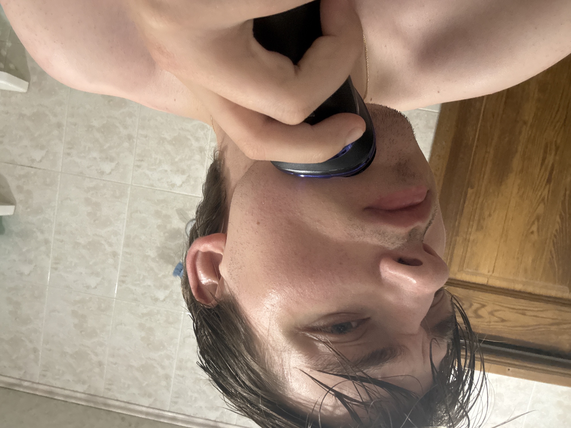 The image shows a person in a bathroom, with tiled walls and a wooden door visible. The person is holding a black electric razor to their face and appears to be shaving. The lighting suggests it is daytime, and the scene is indoors. The person's hair is wet, indicating they may have recently showered. The focus is on the person's face and the razor.