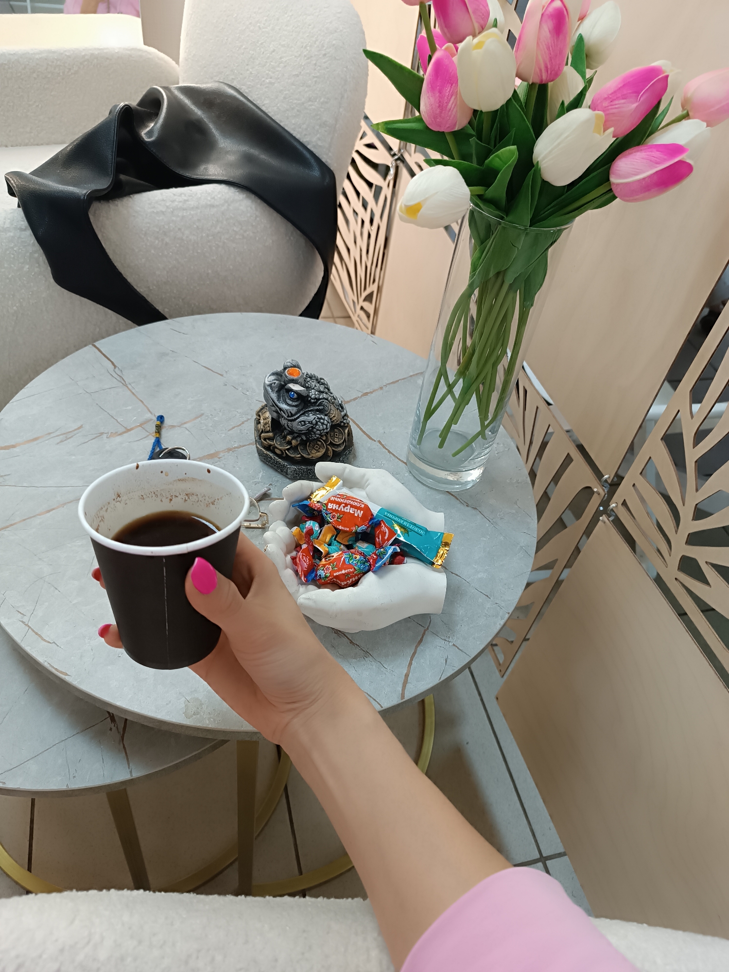 A high-angle shot captures an indoor scene featuring a person holding a black paper cup filled with dark liquid, presumably coffee. The person's hand and part of their forearm are visible, adorned with pink nail polish and clad in a pink long-sleeved garment.
The cup is positioned over a round, marble-patterned table that serves as a resting place for several objects. These include a decorative frog figure, a pair of keys, a pair of glasses, a white hand-shaped bowl filled with colorful wrapped candies, and a clear glass vase containing a bouquet of white and pink tulips.
Behind the table, a white, textured chair with a black accessory is partially visible, along with a decorative wooden screen with leaf-like cutouts. The overall scene presents a cozy, relaxed atmosphere. The location is within a city named Unknown, Unknown.