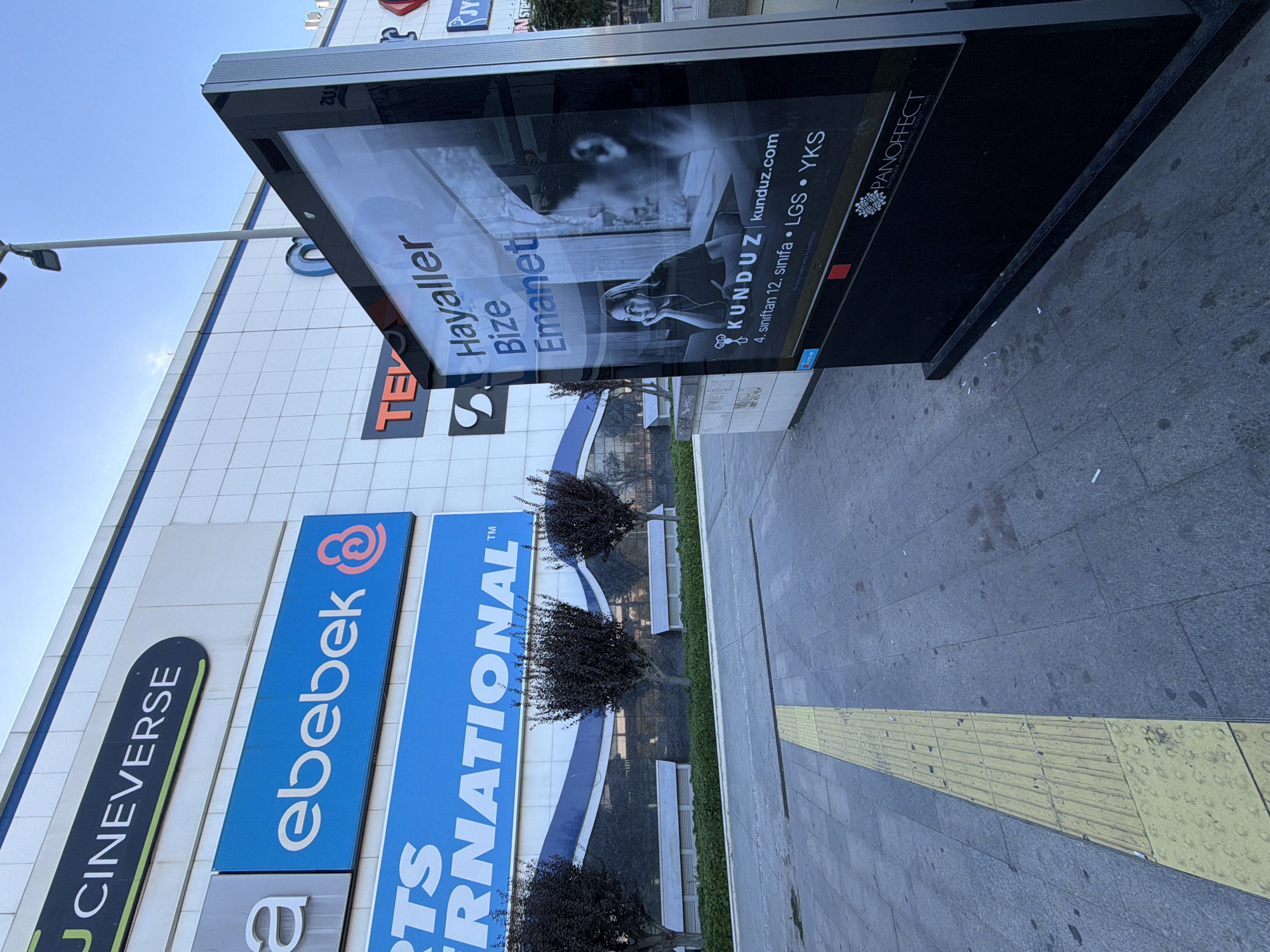 The image shows a shopping mall with several store signs visible. The signs include "JCINEVERSE," "ebebek," and "RTS RNATIONAL." In the foreground, there is a digital advertising display screen showing an advertisement for "KUNDUZ." The screen displays text such as "Hayaller Bize Emanet," "kunduz.com," and educational exam acronyms like "LGS" and "YKS." The digital display is located on a sidewalk with a yellow tactile paving strip. The surrounding environment appears to be an urban setting with a clear sky. This scene is located in Kadıköy, Turkey, a district known for its shopping centers and public spaces.