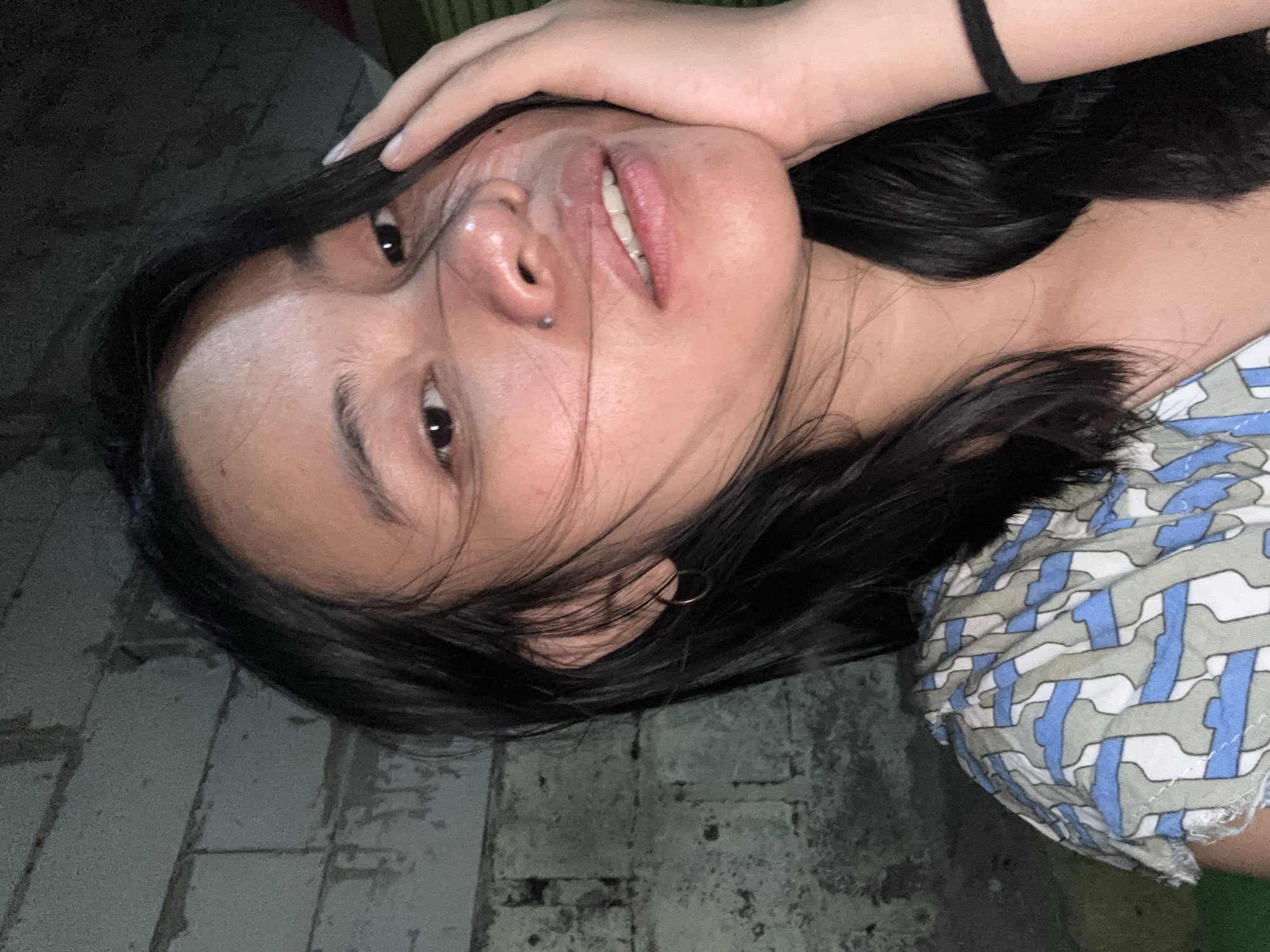A close-up, low-angle, night-time outdoor selfie shows a young woman in East Jakarta, Indonesia. She has dark hair, a small nose piercing, and is wearing a blue and white patterned shirt. Her right hand is supporting her face, and her left hand is visible near her head. The background consists of a weathered brick wall.