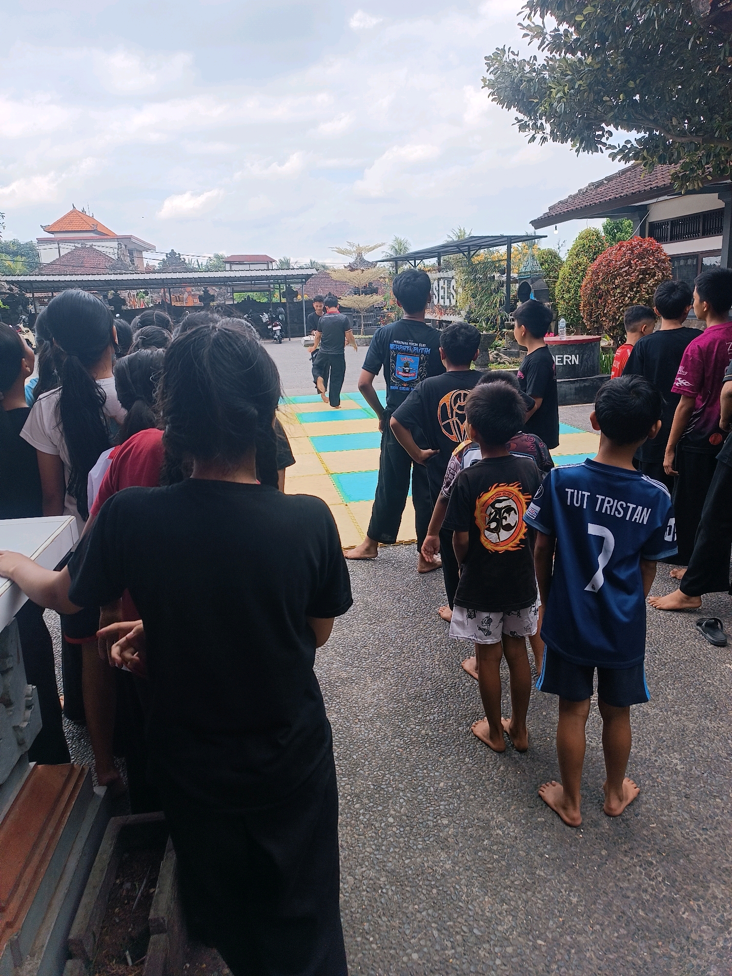 The image shows a group of people, mostly children and teenagers, standing in a line outdoors. They are dressed in casual clothing, with many wearing black t-shirts. Some of the children are barefoot. In the background, there are buildings with traditional Indonesian architecture, including tiled roofs and carved stone elements. There is also a paved area with colorful mats laid out, suggesting an outdoor training or event space. The sky is overcast with clouds. The overall scene suggests a gathering or activity taking place at a cultural or educational facility in Selemadeg, Indonesia.