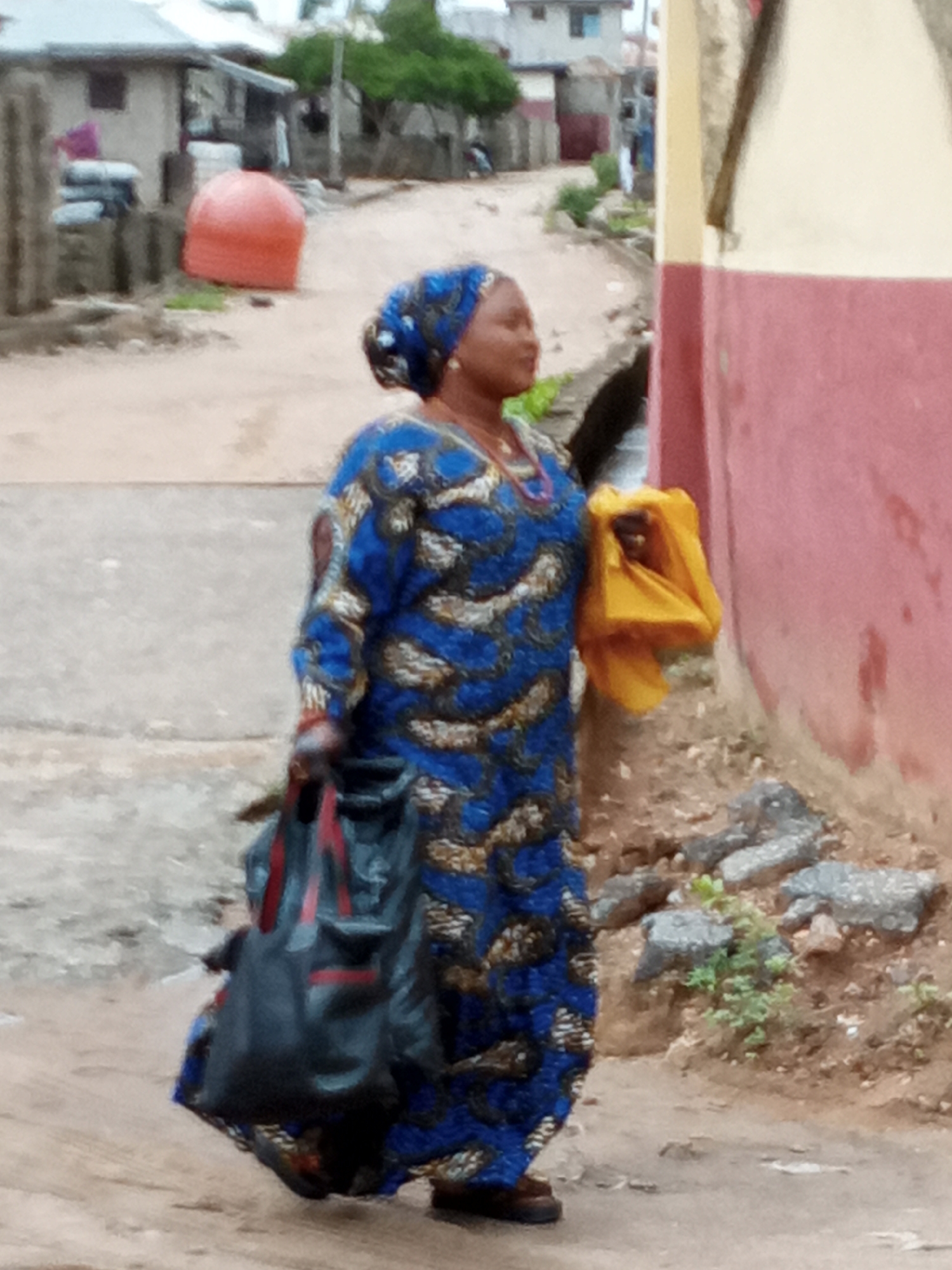 A woman in a blue and gold patterned long dress and headwrap is walking down a dirt road. She is carrying a large black tote bag in one hand and a yellow cloth in the other. The road appears to be in a residential area, with houses and buildings visible in the background. There is a large orange object in the background, possibly a water tank or similar container. The road surface is dirt and there are some rocks and vegetation at the edge of the road. The location is the city of Unknown, Unknown.
