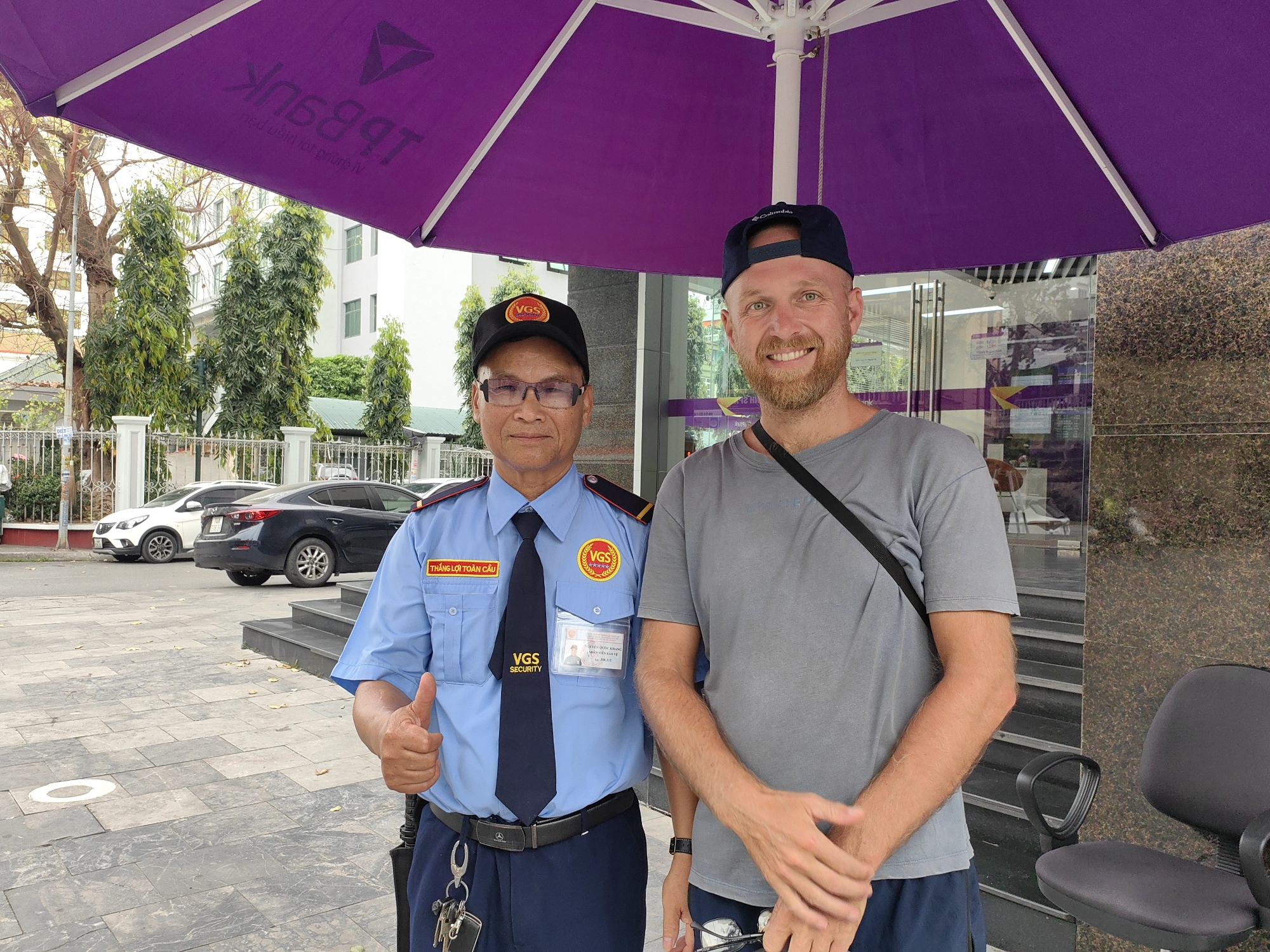 The image shows two men standing under a large purple umbrella. One man is wearing a blue security guard uniform with the name "THANG LOI TOAN CAU" on a name tag, along with patches that say "VGS SECURITY". He has black glasses and a cap with "VGS" on it. He is giving a thumbs-up gesture. The second man is wearing a grey t-shirt, a dark-colored cap, and a black strap across his body. He has a beard and is smiling. Behind them are cars parked on a city street and some trees. Part of a building with glass doors can be seen behind the men. According to metadata the location is Hong Gai Ward, Vietnam.