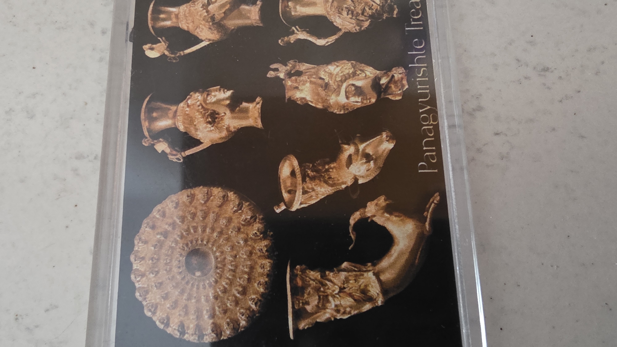 The image shows a postcard or photograph of the Panagyurishte Treasure. The treasure, composed of golden vessels and rhytons, is displayed against a dark background. The objects include a phiale with a Gorgon head, a ram-headed rhyton, a stag-headed rhyton, and other vessels. The text "Panagyurishte Treasure" is visible in the image. The specific location within the city of Blagoevgrad, Bulgaria, is not evident from the image.