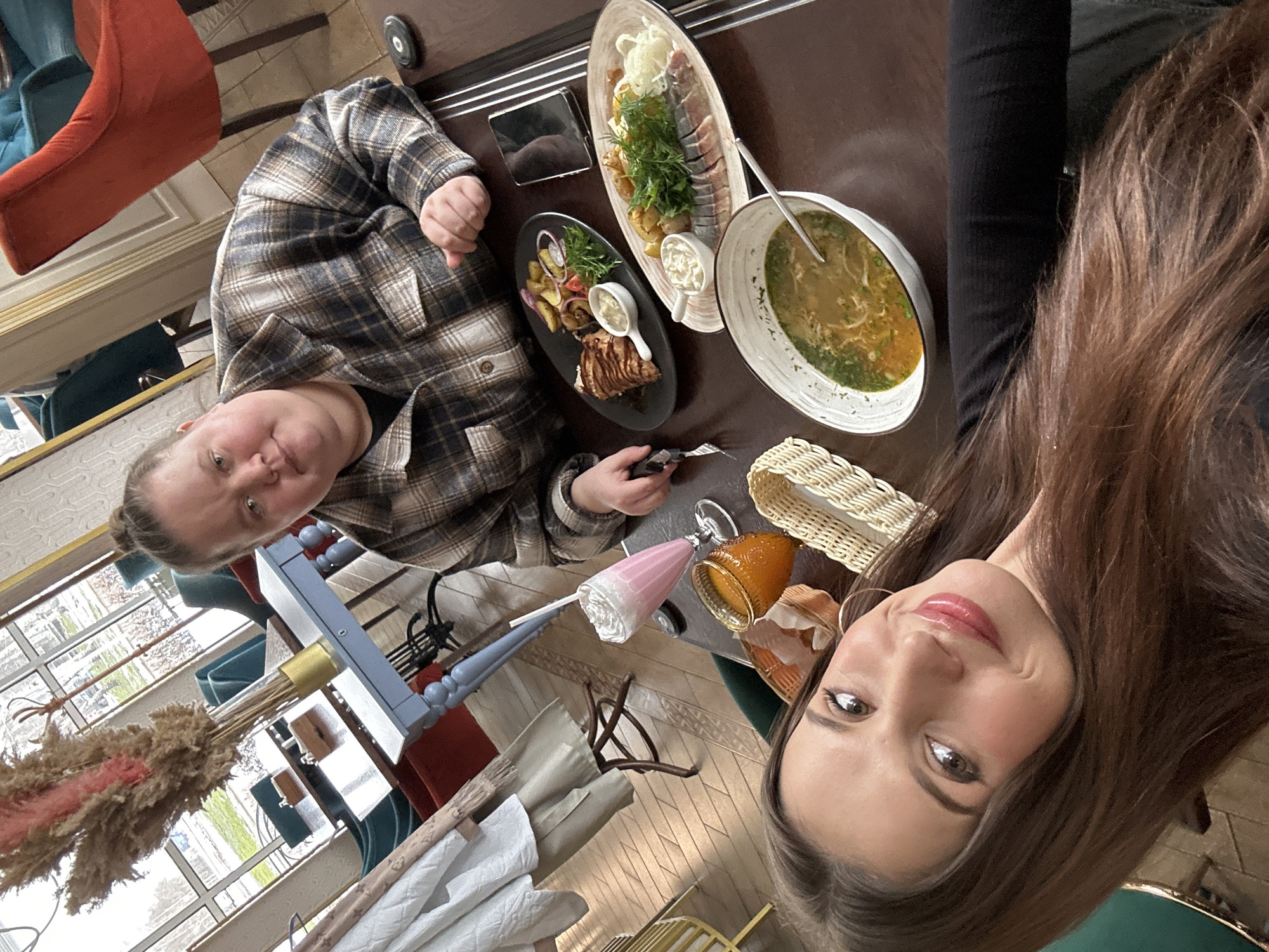 Here is a detailed description of the image:
**Scene:** An indoor setting, presumably a restaurant, based on the food and seating arrangements. The perspective is a selfie, with two people present.
**People:**
* **Person 1:** Located on the left side of the frame, wearing a plaid shirt. Facial features are visible.
* **Person 2:** Positioned on the right side of the frame, with long brown hair. Facial features are clearly visible, suggesting this person is taking the photo.
**Objects:**
* A table filled with food. There is a bowl of soup, a plate with what appears to be meat and potatoes, and a long platter with a variety of items, including fish, greens, and sauces.
* A smartphone on the table.
* A bread basket.
* A pink milkshake.
* There is an umbrella stand in the background.
* Chairs and other restaurant decor are partially visible in the background.
**Actions & Interactions:**
* The image captures a moment of dining and taking a selfie, suggesting a social interaction.
**Environment:**
* The lighting seems natural, possibly from windows, based on the shadows and the light in the background.
* The restaurant has a formal and upscale ambiance.
**Location**
Based on the prompt's information, the location is Chaikovsky, Russia.