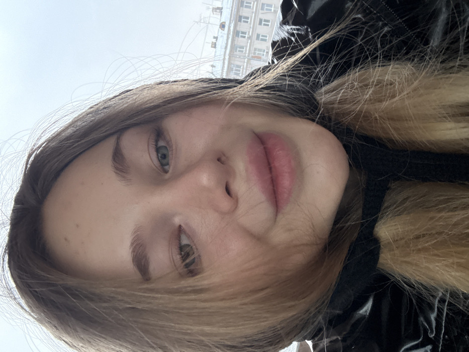 A young woman's face fills the frame, tilted to the side. She has light skin, defined eyebrows, and green eyes. Her lips are pink and full. Her hair is long, blonde with darker roots, and appears slightly damp. She's wearing a black, shiny jacket, possibly with a hood. In the background, blurred buildings suggest an urban setting, possibly during overcast weather. The overall impression is casual and informal.