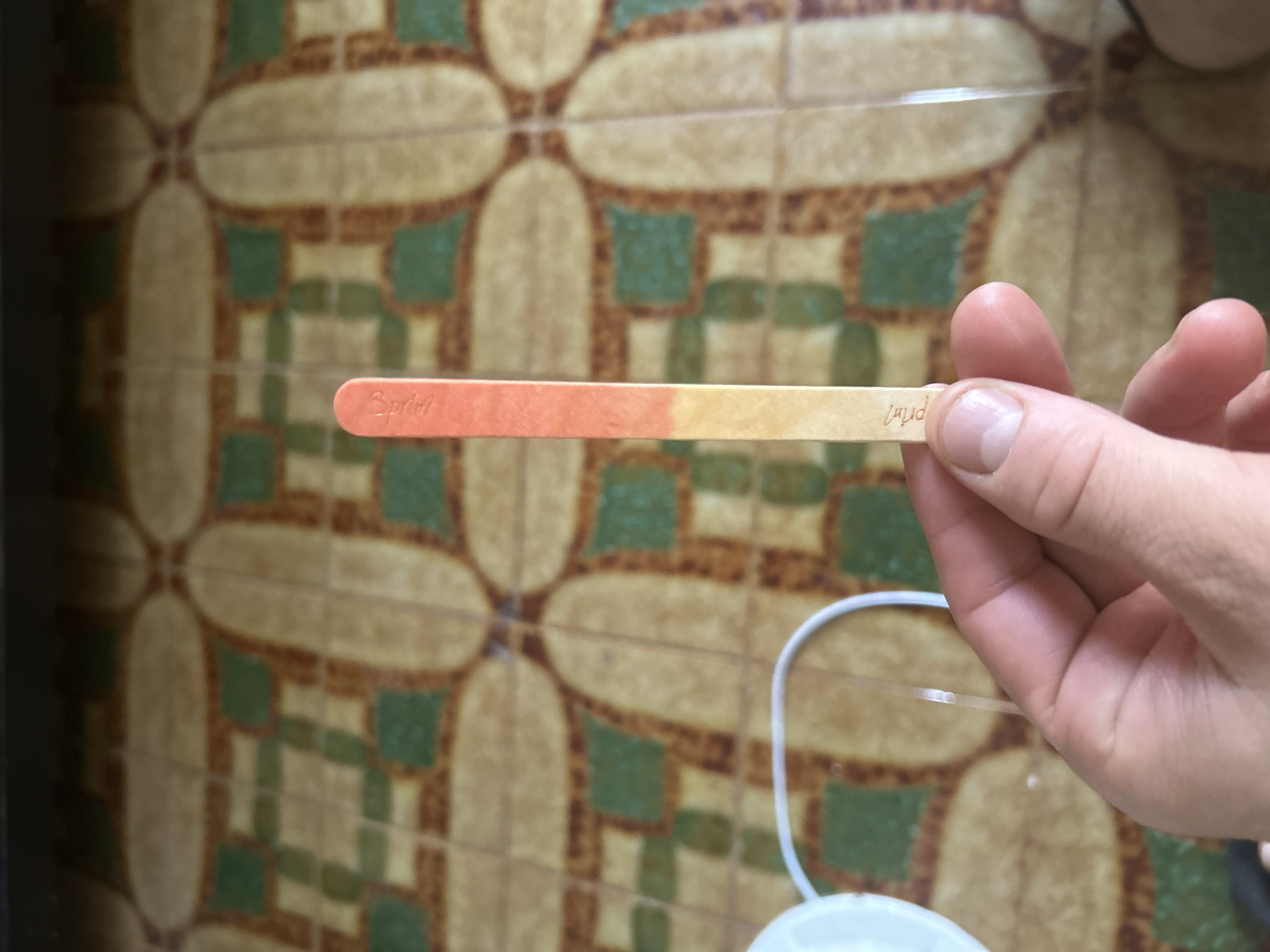 The image shows a close-up of a hand holding a litmus paper strip. The litmus paper strip is a light-colored wooden popsicle stick with a colored strip running along its length. The strip transitions in color from a deep orange on the left to a pale yellow on the right, suggesting a measurement of pH. The hand holding the stick has manicured fingernails, and the skin texture is visible.
In the background, there is a tiled surface with a repeating pattern of beige oval shapes surrounded by green and brown squares. This pattern suggests an indoor setting, possibly a bathroom or kitchen, with decorative tiles. A white cord, possibly from an electronic device, is also visible in the background, leading down out of frame.
The lighting in the image appears to be artificial, casting a warm glow on the scene. There are no explicit indicators of time of day or weather conditions. The overall impression is a moment of measurement or testing, possibly related to water quality or a science experiment. The visible text on the litmus paper is too blurry to read clearly, but it appears to be a brand name or indication of what is being tested. The location context is identified as Barcelona, Spain.