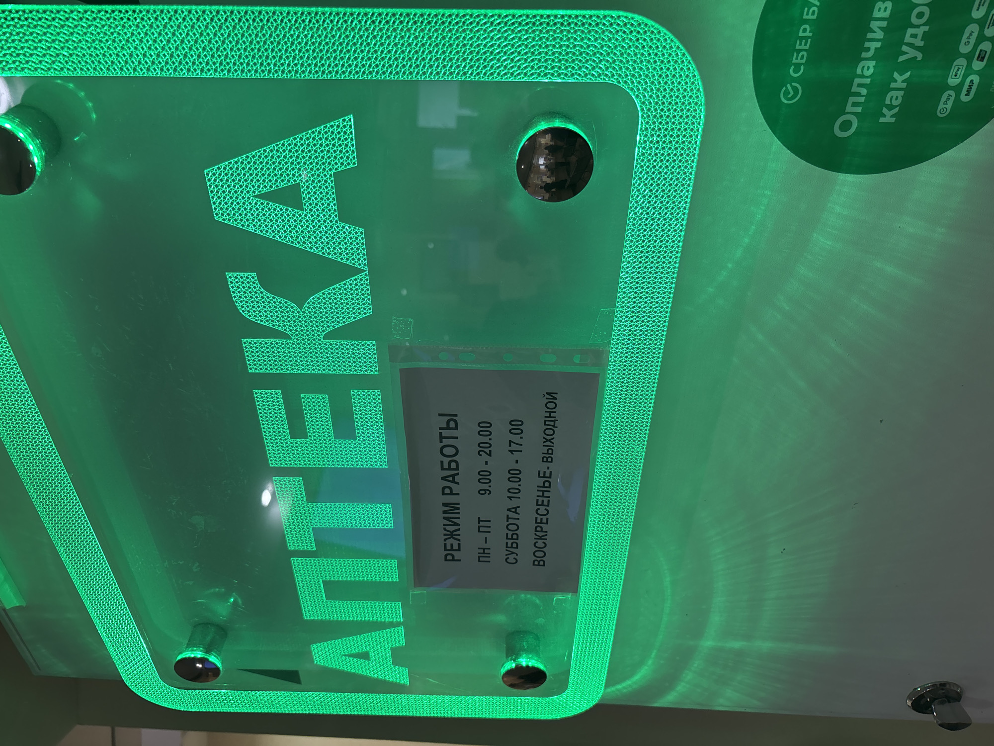 Here's a concise description of the image:
The image is a close-up shot of a bright green sign for a pharmacy ("АПТЕКА") in Saint Petersburg, Russia. The sign also includes the pharmacy's hours of operation ("РЕЖИМ РАБОТЫ") in Russian: Monday-Friday 9:00-20:00, Saturday 10:00-17:00, Sunday - closed ("ВОСКРЕСЕНЬЕ- ВЫХОДНОЙ"). In the upper right corner of the image there is part of another sign which mentions the words "СБЕР БА" ("Sber Bank") and the ways to pay for purchases.