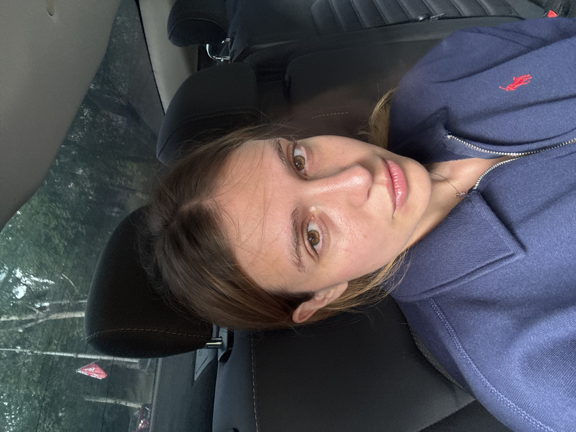 A woman with fair skin, brown hair, and light brown eyes is lying in the backseat of a car. She is wearing a navy blue jacket with a red logo on the chest. The car's seat and headrest are black. The window shows trees and a red triangular sign outside. Based on the style of architecture and vegetation visible through the window, the location is estimated to be Tbilisi, Georgia.