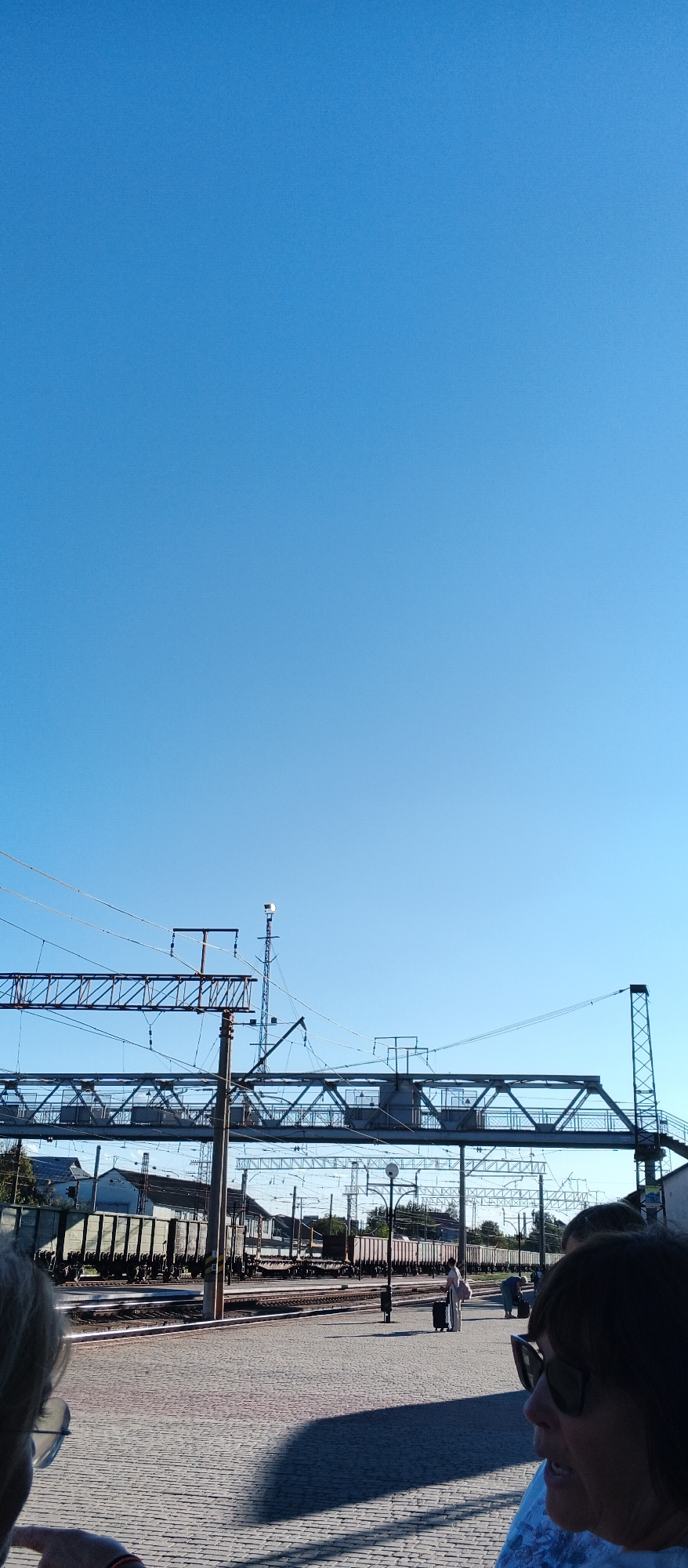 The image captures a view of a train station in Mukachevo, Ukraine, under a clear blue sky. In the foreground, the heads and shoulders of two individuals are visible, partially obscuring the scene beyond. The individuals appear to be facing away from the camera and looking towards the train tracks.
The midground of the image features a train station platform paved with cobblestones. Several train cars are parked on the tracks, some appearing to be freight cars. Overhead, a network of power lines and a pedestrian bridge or overpass can be seen, supported by metal structures and poles. A tall communication tower with an antenna is also visible against the sky.
In the background, various buildings and trees are present, suggesting the urban environment surrounding the train station. The overall atmosphere is one of a typical day at a train station, with people waiting and trains in transit. The lighting suggests it is daytime, likely morning or afternoon, with the sun casting shadows on the platform.