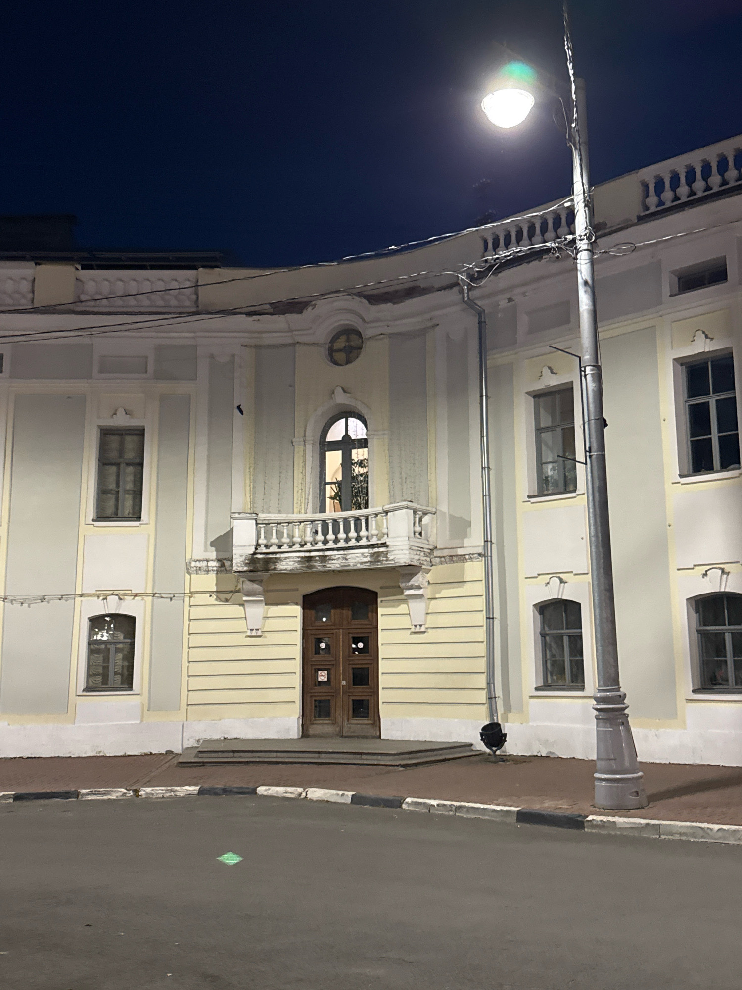 The image captures a building facade at night in Tver, Russia, illuminated by a streetlamp. The building is a light yellow color with white architectural detailing, featuring multiple windows of varying sizes and shapes. A prominent feature is a balcony with a white balustrade above a set of double wooden doors. A lamppost stands in front of the building, its light casting a glow on the scene. The ground in front of the building is paved with asphalt and gravel. The sky above is a deep blue, indicating the late hour. The overall scene is one of quiet urban nighttime ambiance.