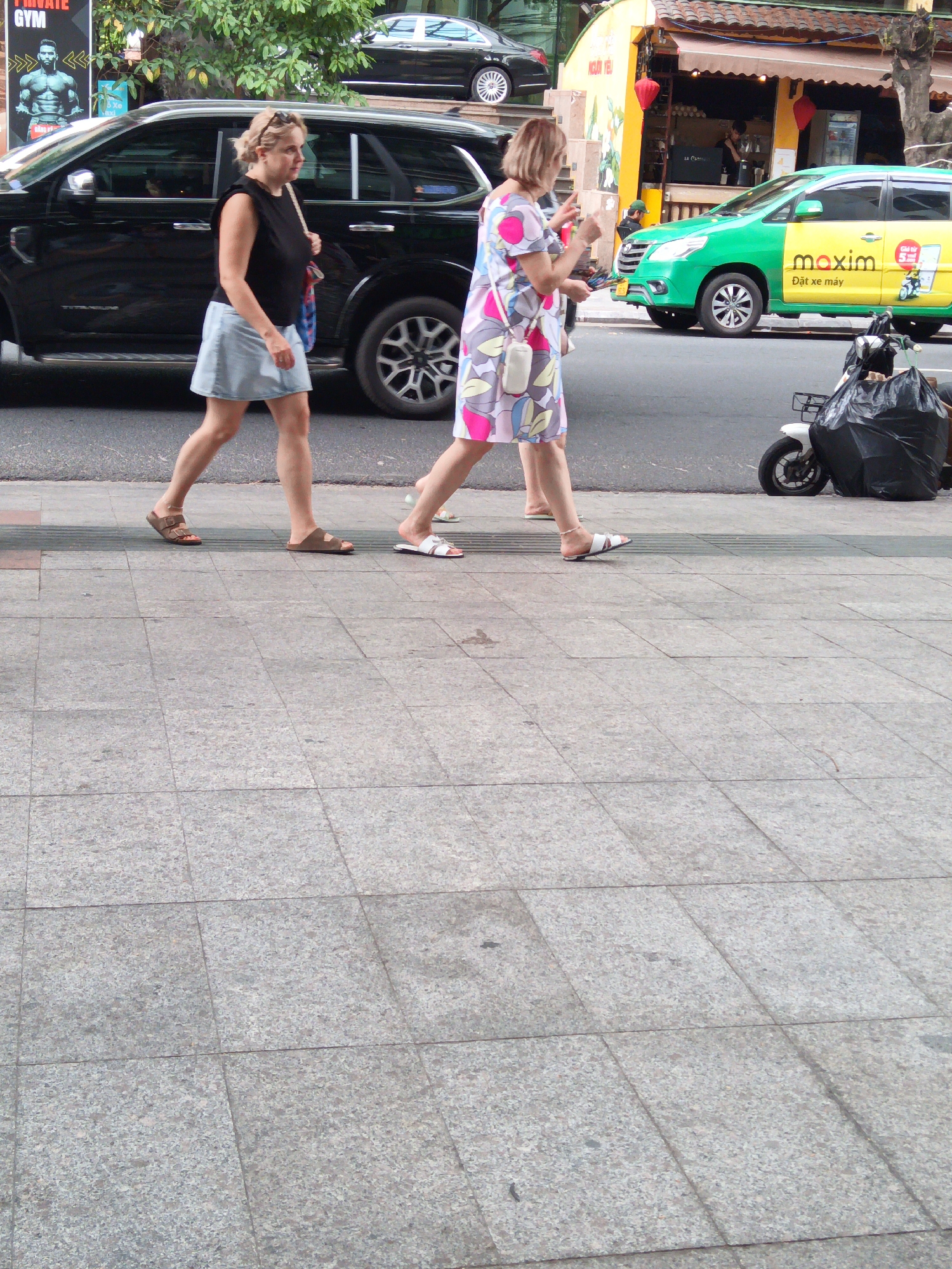 In Nha Trang, Vietnam, two women walk along a paved sidewalk. The woman on the left, with blonde hair pulled back, wears a black sleeveless top and a light blue denim skirt. She has on brown Birkenstock-style sandals. The woman on the right, also with blonde hair, is dressed in a colorful floral print dress with short sleeves and white sandals with a strap. A white crossbody bag is slung over her shoulder.
Behind them, a black SUV is parked at the curb. To the right of the women, a green and yellow taxi with "maxim" branding is stopped, its front passenger door open. A white scooter with a large black trash bag strapped to its rear is parked next to the taxi. A yellow building with awnings and red lanterns visible is behind the taxi, suggesting it is a commercial area, likely a cafe or shop. The background shows a glimpse of trees and another vehicle, a dark-colored car, further down the street. The scene is illuminated by daylight, casting shadows on the sidewalk.