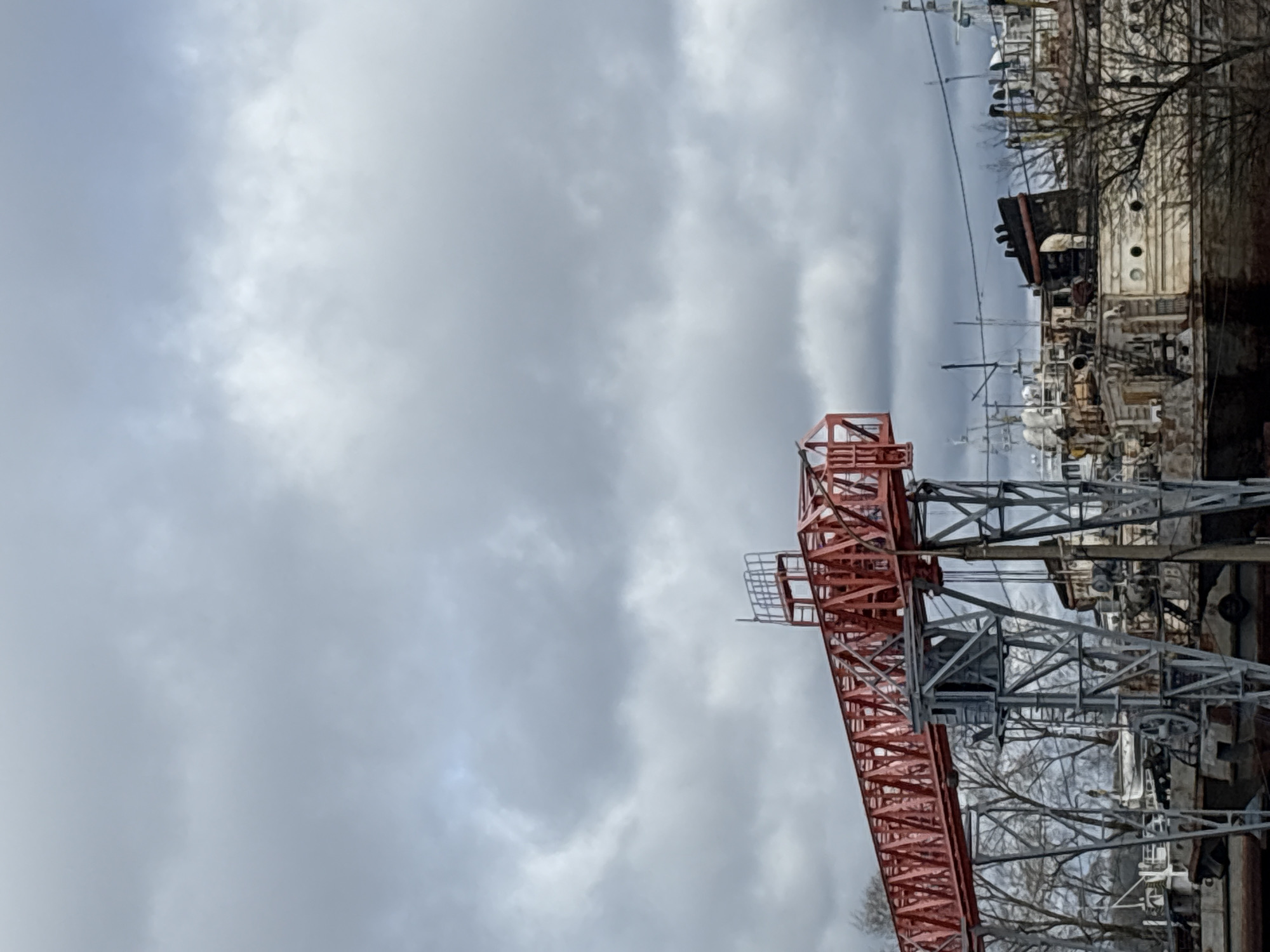 The image shows a gray cloudy sky above an industrial area with a red and gray metal crane in the foreground. In the background, there are numerous boats and ships docked, as well as tree branches and other industrial structures. Based on available information and visual cues, this image likely shows a section of the boat repair dock in Chaikovsky, Russia.