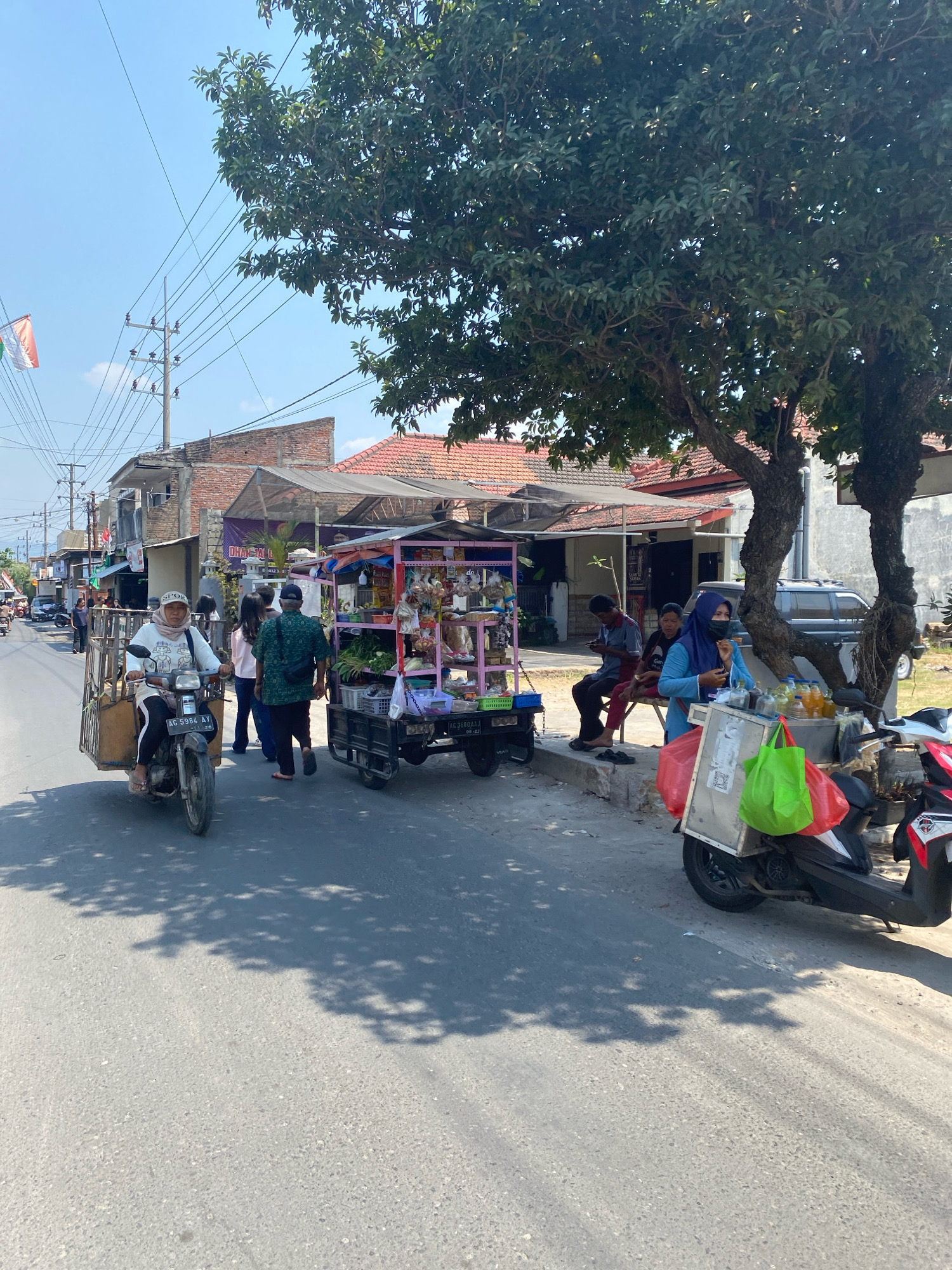 The image depicts a street scene in the city of Kediri, Indonesia, likely during the daytime under clear skies. A paved road runs through the center of the frame, bordered by buildings and trees. On the left side of the road, a person wearing a headscarf and a white shirt is riding a motorcycle with a sidecar. Further down the road, several pedestrians are walking, and various shops and stalls are visible.
In the foreground, a colorful cart selling goods, possibly food or produce, is parked on the right side of the street. Next to the cart, a motorcycle is parked, adorned with several bags, including a red and a green one. A person in a blue hijab and mask is standing near the motorcycle, holding what appears to be a phone. Two men are seated on a bench or low wall to the left of the cart.
The environment suggests a bustling commercial area with a mix of residential and business structures. Overhead power lines crisscross the sky, and shadows from trees cast patterns on the road, indicating sunlight filtering through foliage. The overall impression is that of a typical urban street in Indonesia, characterized by local transportation, street vendors, and everyday activities.