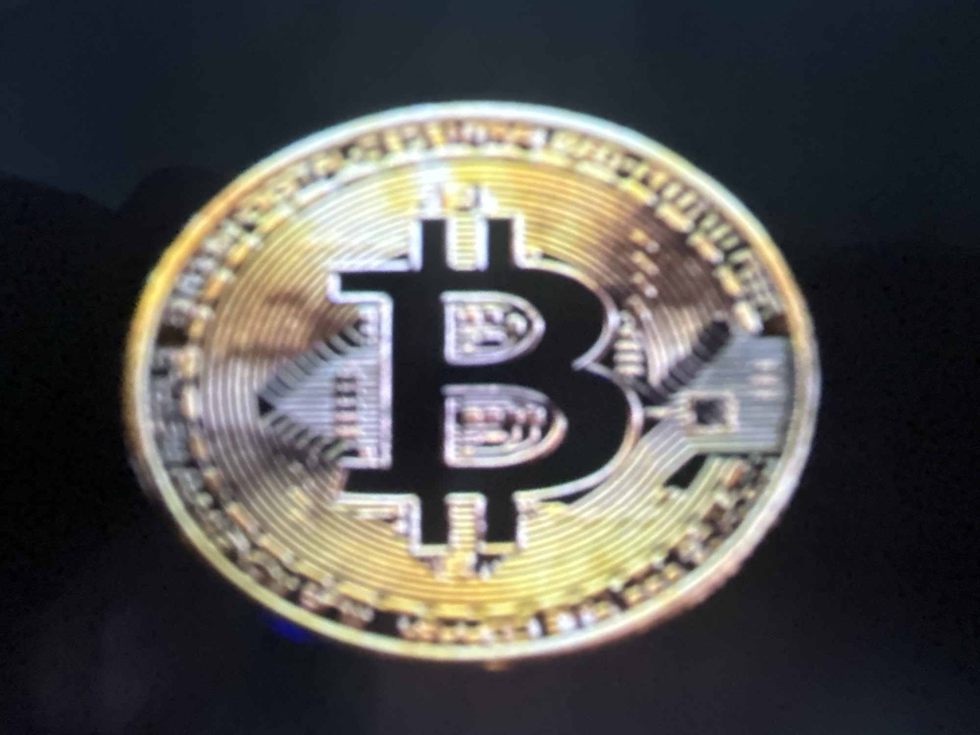 This is a close-up, slightly blurry photograph of a gold-colored Bitcoin cryptocurrency coin. The coin is the sole subject, positioned centrally against a dark, out-of-focus background. The obverse of the coin is visible, featuring the iconic Bitcoin symbol, a stylized "B" with vertical lines. The edge of the coin has intricate detailing, with radial patterns and what appear to be repeating text or designs, though these are not legible due to the blurriness. The lighting creates a metallic sheen on the coin, highlighting its gold tone. There are no people, discernible setting, or time-of-day cues visible. The image appears to be a still shot of the coin, with no activity or event taking place.