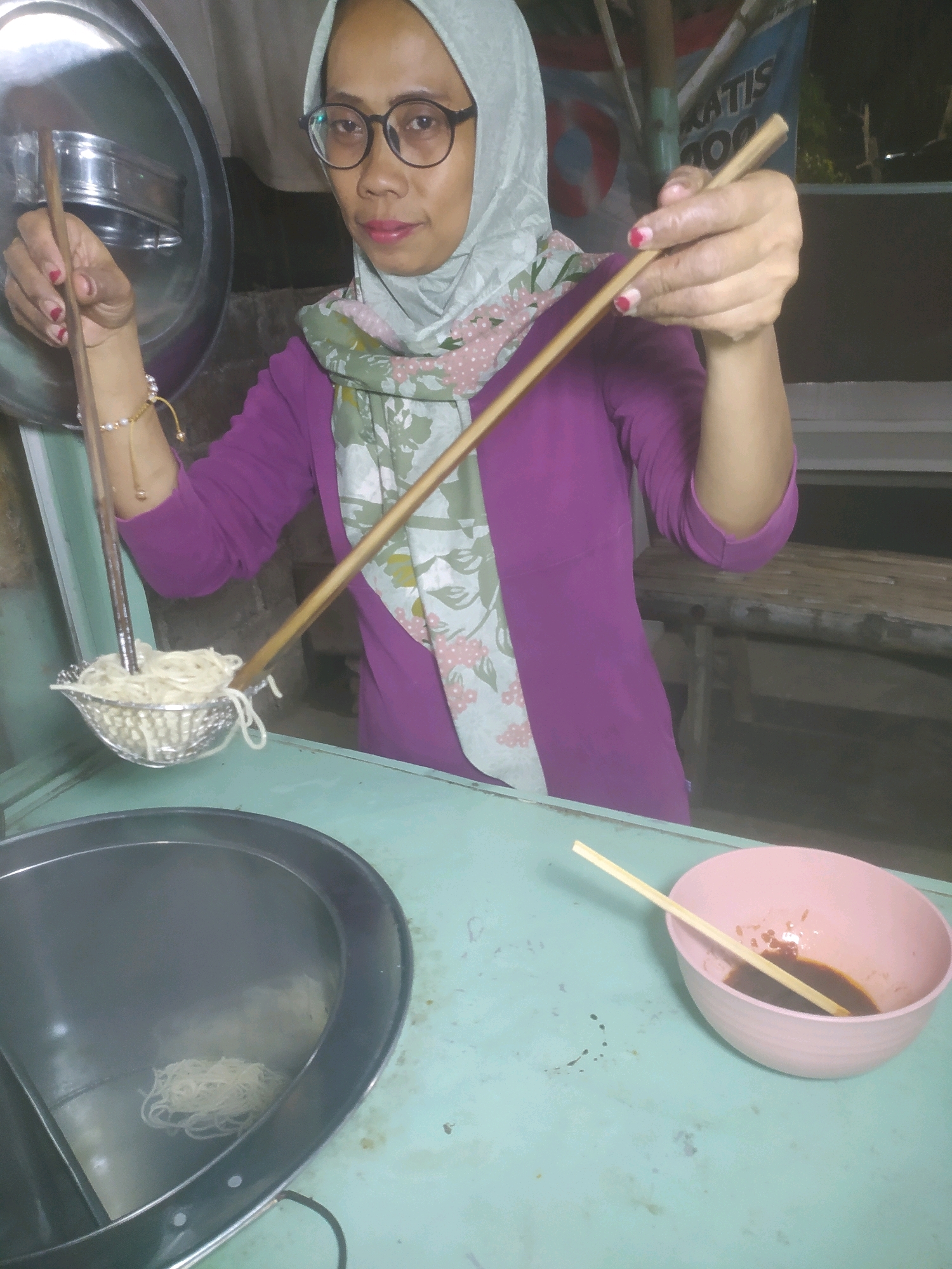 A woman wearing a hijab and glasses is preparing noodles in what appears to be a street food stall in Karanggayam, Indonesia. She holds a metal strainer filled with noodles in her left hand, while her right hand holds a pair of chopsticks over the strainer. A bowl with dark liquid, presumably sauce or broth, and another pair of chopsticks rest on a turquoise counter in the foreground. A larger metal pot or basin is visible to the left of the woman. The background suggests an outdoor or semi-outdoor setting with wooden structures and a banner with text, including "GRATIS". The lighting is dim, indicating it might be evening or night.