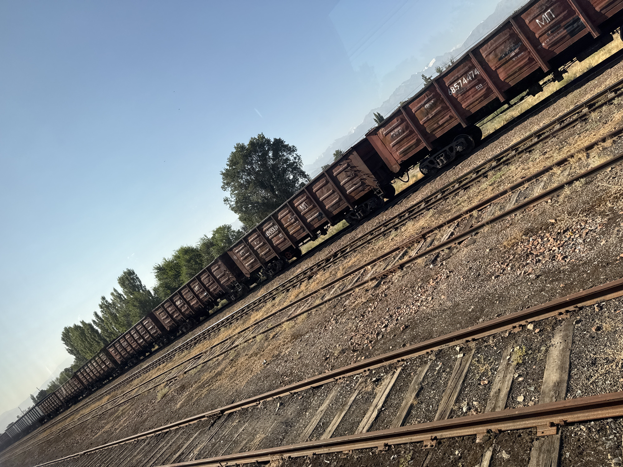 The image shows a train with many open-top cargo cars on a railway track in Ivanovka, Kyrgyzstan. The train cars are rusted brown and have markings and numbers on their sides. The railway tracks are made of steel and rest on wooden sleepers, with gravel and dirt ballast. The scene is viewed from an elevated perspective, looking down the length of the train. On the right side of the train, there are green trees and in the background, there are mountains under a clear blue sky. The sun is shining, casting shadows and illuminating the scene. There are no people visible in the image. The overall impression is of an industrial or freight transportation setting in a natural landscape.