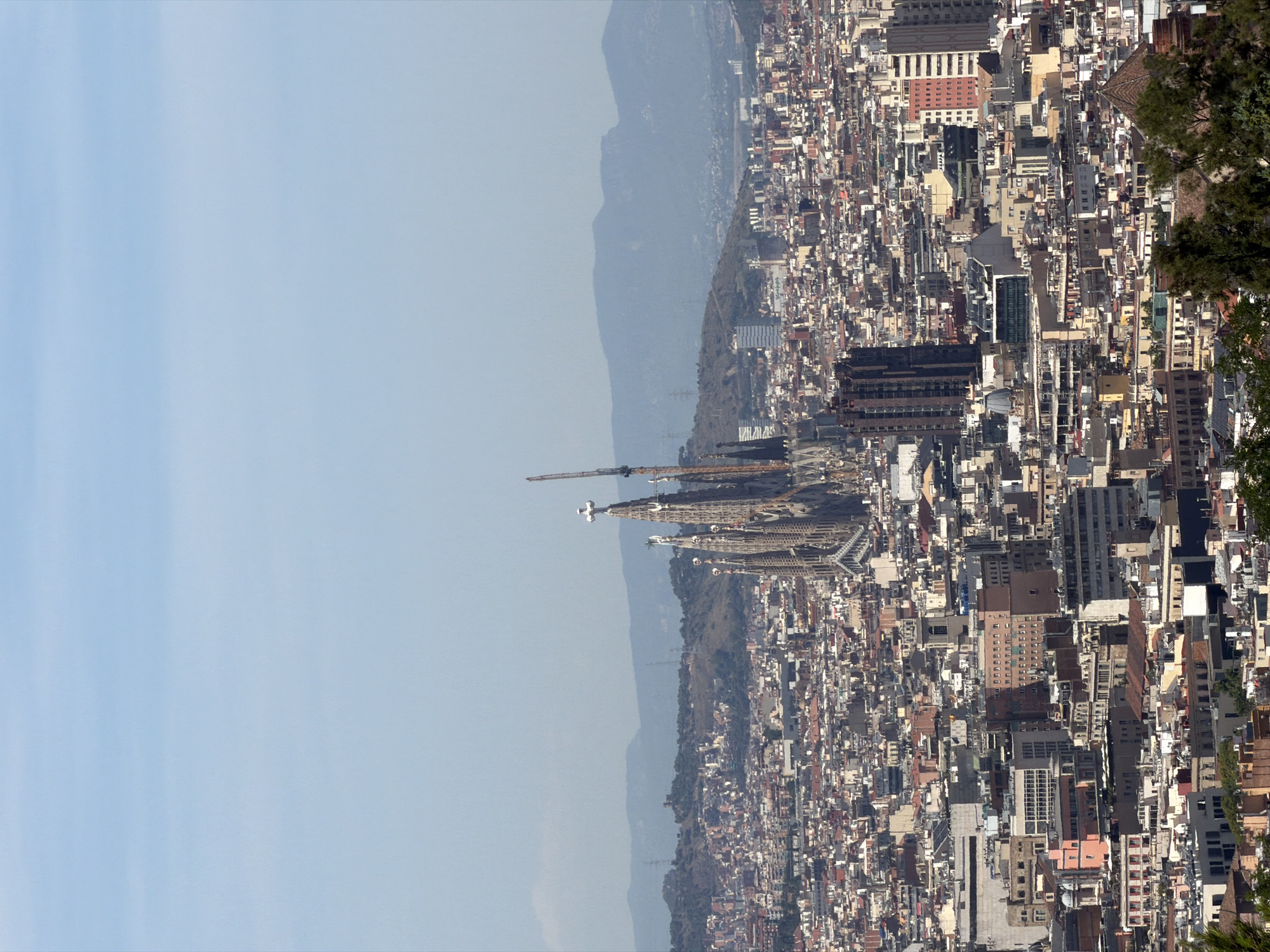 This is a high-angle, wide shot of the city of Barcelona, Spain, taken on a clear, sunny day. The Sagrada Familia, a towering basilica with intricate spires and a prominent cross at its apex, dominates the mid-ground on the left side of the frame. Construction cranes are visible near the basilica, indicating ongoing work. The foreground and mid-ground are filled with a dense urban landscape of buildings of various sizes and architectural styles, characterized by terracotta roofs and a mix of modern and older structures. Beyond the city, rolling hills and distant mountains form the backdrop, under a pale blue sky. There are no people or animals clearly visible in the image. The scene captures a panoramic view of a bustling metropolis under a bright sky.